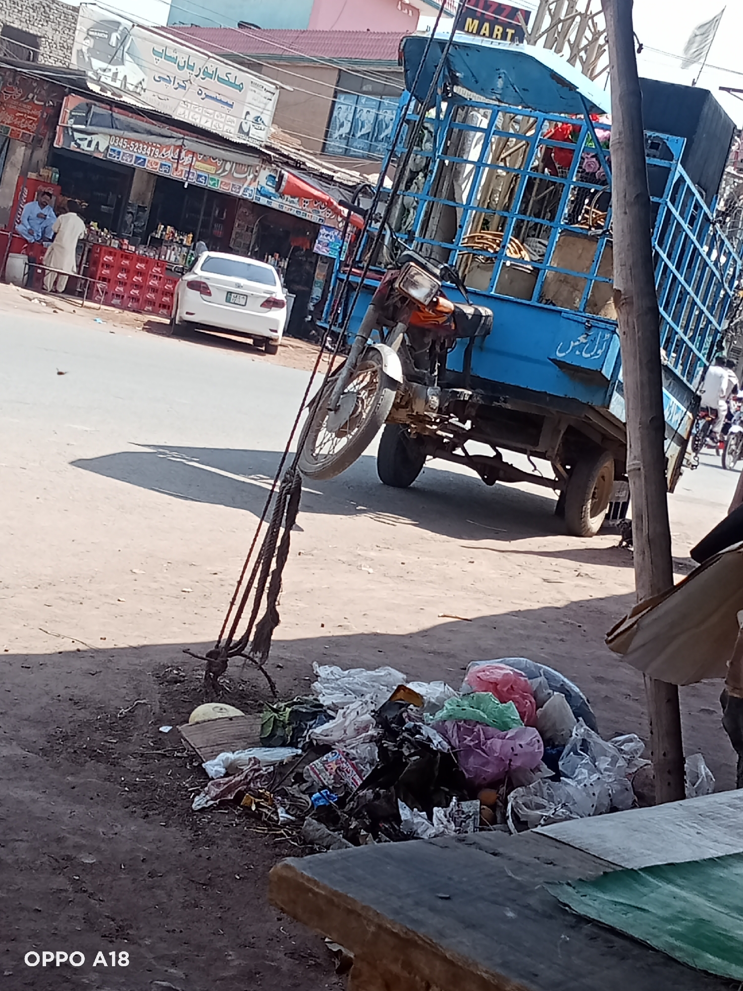 The image captures a bustling street scene, likely in an urban or semi-urban area. In the immediate foreground, a weathered wooden pole stands prominently, secured with thick ropes, below which lies a significant pile of trash consisting of various plastic bags, cardboard, and other debris on a dirt patch. A wooden surface, possibly a table or counter, is partially visible at the bottom right. The bottom left corner shows an "OPPO A18" watermark.
In the midground, a paved road stretches across the frame. On the left side of the road, a white sedan car is parked. Further left, two men are visible outside a storefront; one is seated on a low barrier, and the other stands nearby, both wearing traditional attire. Red plastic crates are stacked in front of this shop. Towards the center, a motorcycle is prominently featured, appearing to be leaning against the pole or part of a larger three-wheeled vehicle, with its front wheel elevated.
On the right side of the road, a blue three-wheeled cargo vehicle, with a caged back, is moving away from the viewer. Another person on a motorcycle is visible further down the road behind it. Long shadows cast by the motorcycle and pole on the road indicate a sunny day, likely during the morning or late afternoon.
In the background, a row of storefronts lines the street. A large white banner above one shop displays text in Urdu/Arabic script, which reads "ملک انور پان شاپ" (Malik Anwar Paan Shop) and "پنسره کراچی" (Pansara Karachi). Another sign further back partially shows the word "MART." Below the main banner, a smaller sign includes a phone number: "0345-5233479." The buildings behind the shops feature different colored roofs, including red and pink.