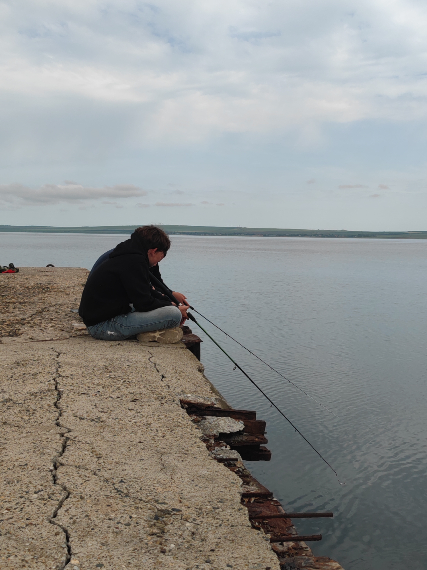 Two people are squatting on a concrete pier in Sennoy, Russia, fishing in a large body of water. The pier is cracked and damaged, with exposed rebar along the edge. The people are facing the water, holding fishing rods. The sky is cloudy and the water is calm. A strip of land is visible across the water in the distance.