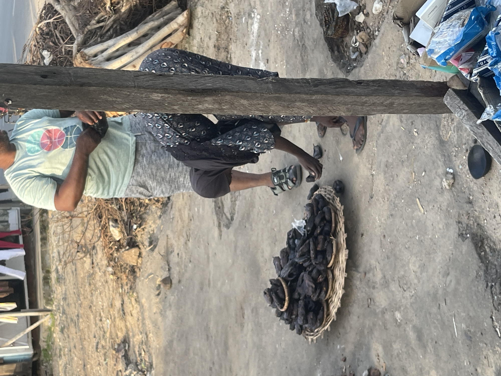 In an outdoor, dusty environment, a woman dressed in a dark, patterned dress and a black headscarf is bent over a pile of dark, dried food items displayed on a woven mat and a smaller woven tray. She is using her hands to sort or select the items. To her left, a man wearing a light green t-shirt with a graphic design and grey shorts stands holding a dark object in his hands. A rough, weathered wooden post stands prominently in the foreground, slightly to the right of center. In the background, there is a structure with hanging clothing, dry brushwood, and scattered debris. The ground is a mixture of dirt and concrete.