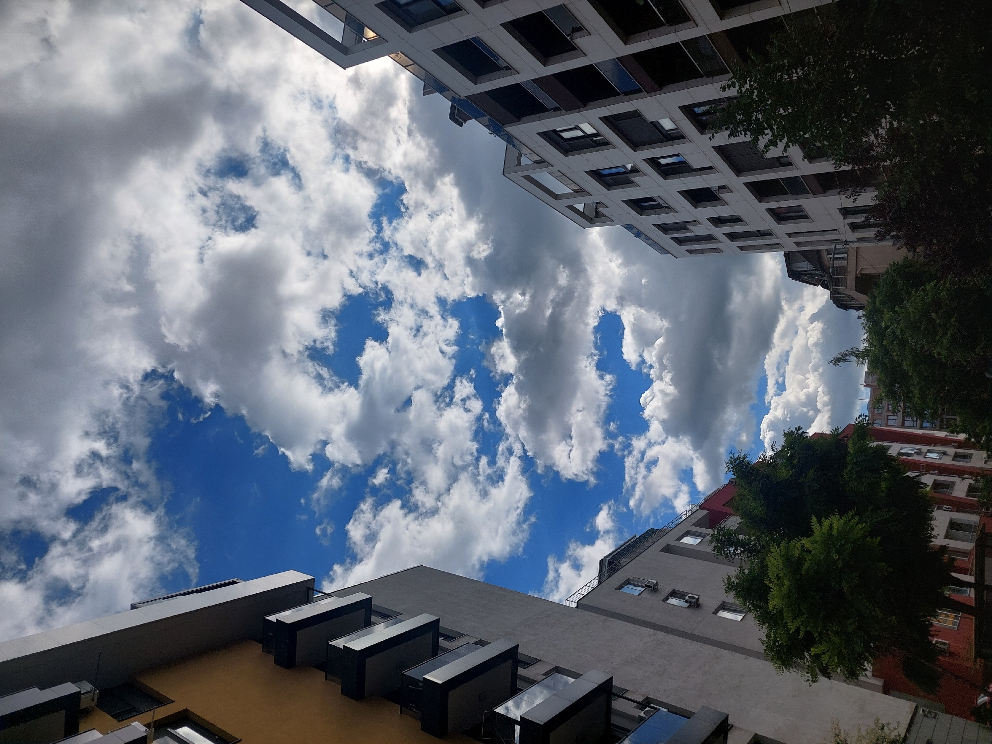The image shows a view looking up at modern apartment buildings against a dramatic sky filled with white and grey clouds. The buildings are contemporary in design, with multiple stories, balconies, and large windows. On the left, a building with a yellow facade and dark balconies is prominent. To the right, a white building with a grid-like pattern of windows and balconies stands out. Trees are visible at the base of the buildings, with lush green foliage. The sky is a mix of bright blue patches and heavy cloud cover, suggesting dynamic weather. The perspective is from below, emphasizing the height of the structures and the vastness of the sky. The scene appears to be in an urban environment, possibly a residential area. This image was taken in Sofia, Bulgaria, on May 15, 2023.