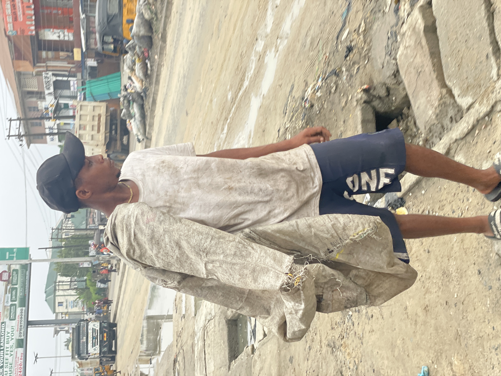 A young male, appearing to be in his late teens or early twenties, with dark skin, stands on a street in Ado, Nigeria. He is wearing a black baseball cap, a soiled white short-sleeved t-shirt, dark blue shorts with a white "ONE" graphic on the left leg, and black-and-white striped slide sandals. A gold-colored chain is visible around his neck. He carries a large, tattered, light-brown woven sack over his left shoulder. His gaze is directed towards the right side of the frame.
The immediate foreground features a broken, uneven concrete walkway with an open drainage gully containing standing water. The street itself is unpaved, muddy, and wet, with visible puddles. In the mid-ground, a white dump truck is parked on the left side of the road. On the right, a second white truck is visible, alongside a pile of green corrugated metal sheets and a partially obscured yellow commercial tricycle. Further down the street on the right, large piles of bagged refuse are accumulated along the roadside.
The background consists of various low-rise commercial and residential buildings, some with corrugated metal roofs. A prominent billboard on the upper left advertises housing, displaying phrases like "U SEF FIT BUY HOUSE... YES, YOU!", "N30,000", "2400/Day", and "Contact Us 09166709090". Several blurry figures are visible in the distance, suggesting pedestrian movement. The overall scene depicts a functional but infrastructurally challenged urban environment.