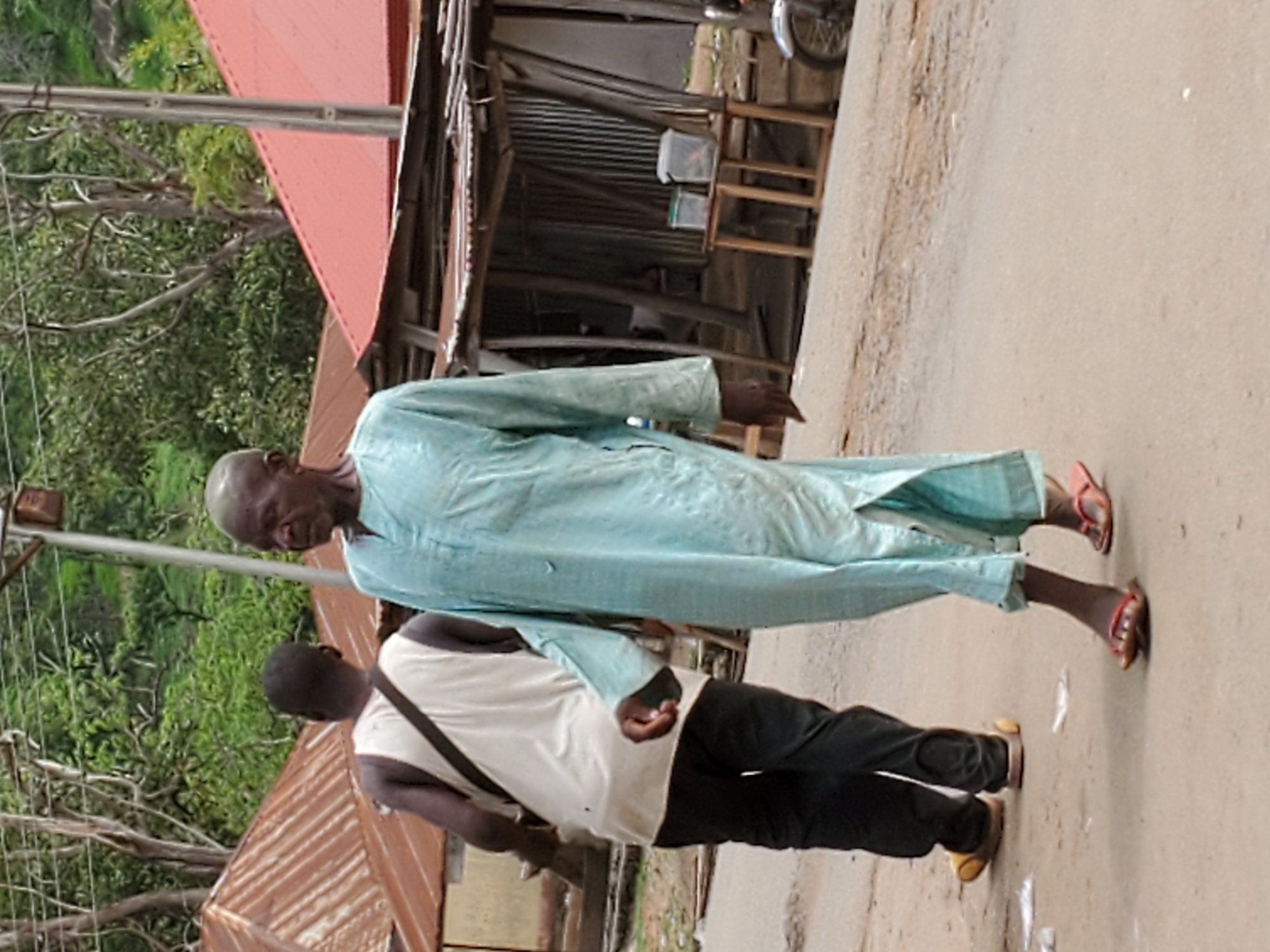 In Mararaba, Nigeria, an older man in a light blue robe and sandals walks along a street next to a younger man wearing a white shirt, black pants, and sandals. The older man appears to be the focus of the younger man’s attention. The setting includes buildings with corrugated metal roofs and trees in the background.