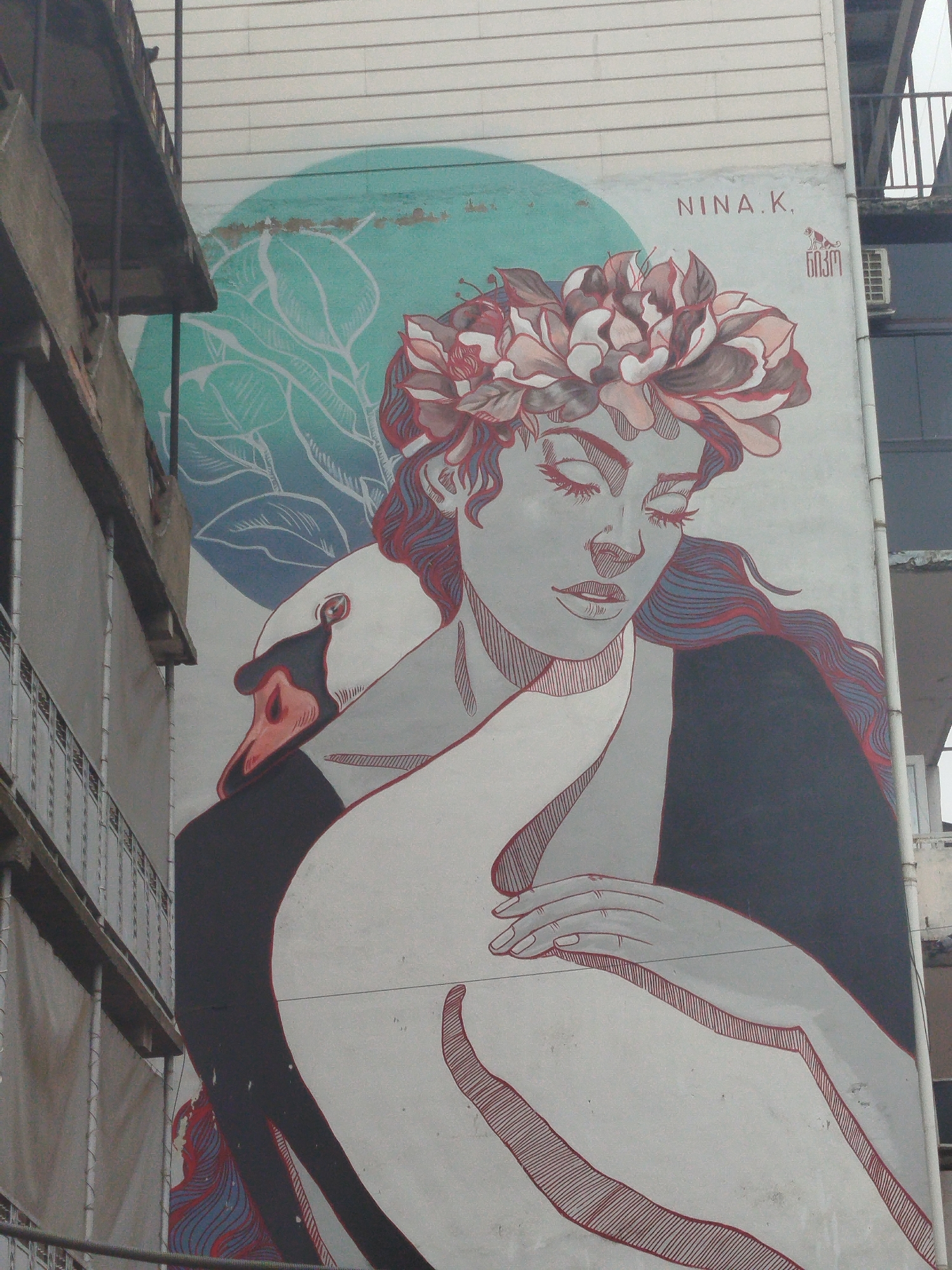 A mural is painted on the side of a building. The mural depicts a woman with a flower crown, her eyes closed, looking down. A swan's head and neck are positioned behind her, with the swan's beak near the woman's face. The mural is rendered in shades of gray, white, black, and red, with a teal background featuring a stylized plant. The text "NINA.K." is visible in the upper right corner of the mural, above a small, stylized animal figure. The building appears to be multi-story, with balconies visible on the left side of the image. The mural is located in Batumi, Georgia.