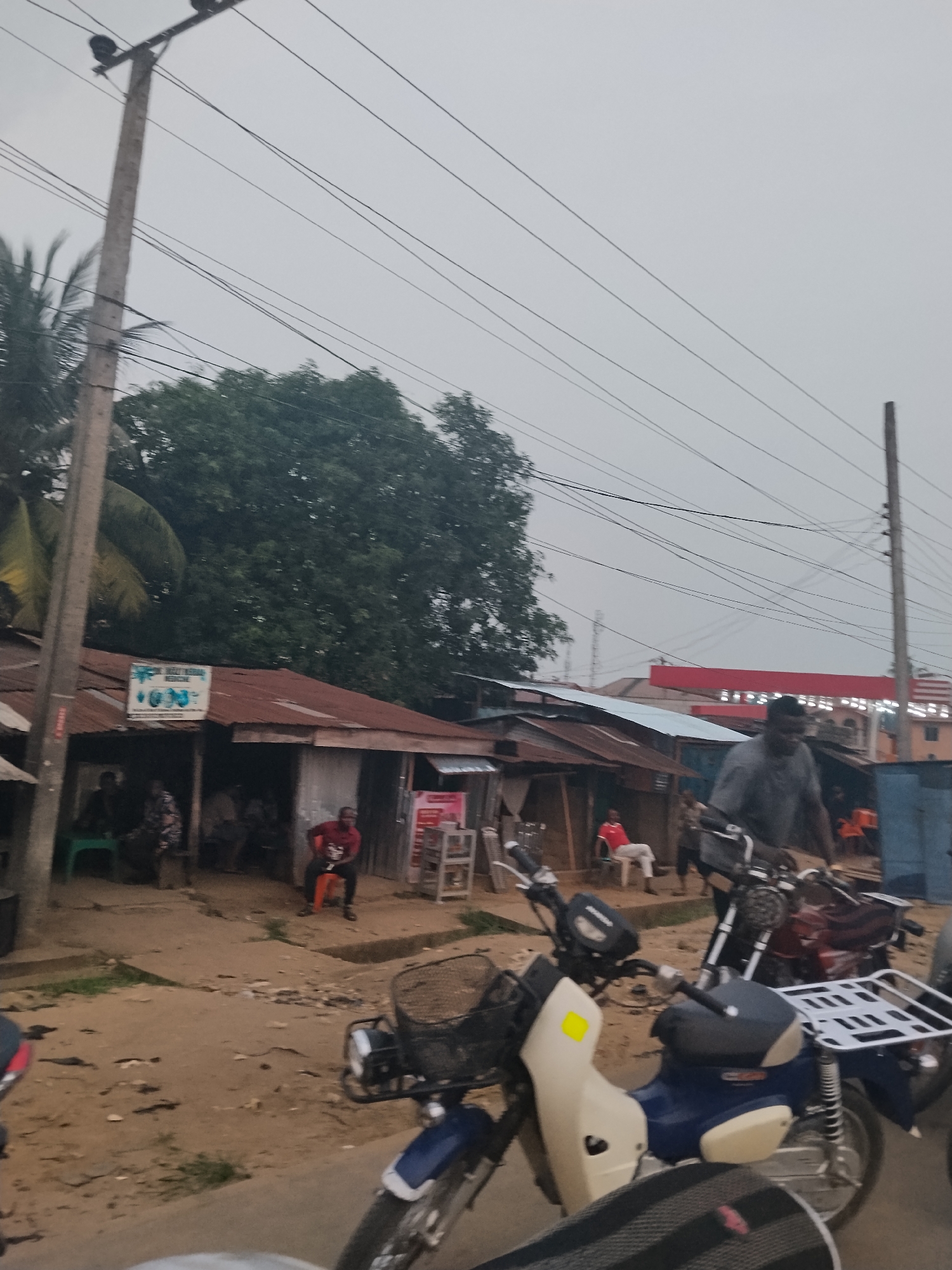 The image captures a street scene in Ura Nta Umuarandu, Nigeria. The foreground features two motorcycles, one primarily blue and white with a front basket and the other maroon and silver. Several people are seen in the background. Some are seated in front of small shops or dwellings with corrugated metal roofs. One man is riding a motorcycle. Overhead, power lines stretch across the sky. The environment appears to be a dirt road with some greenery. The sky is overcast.