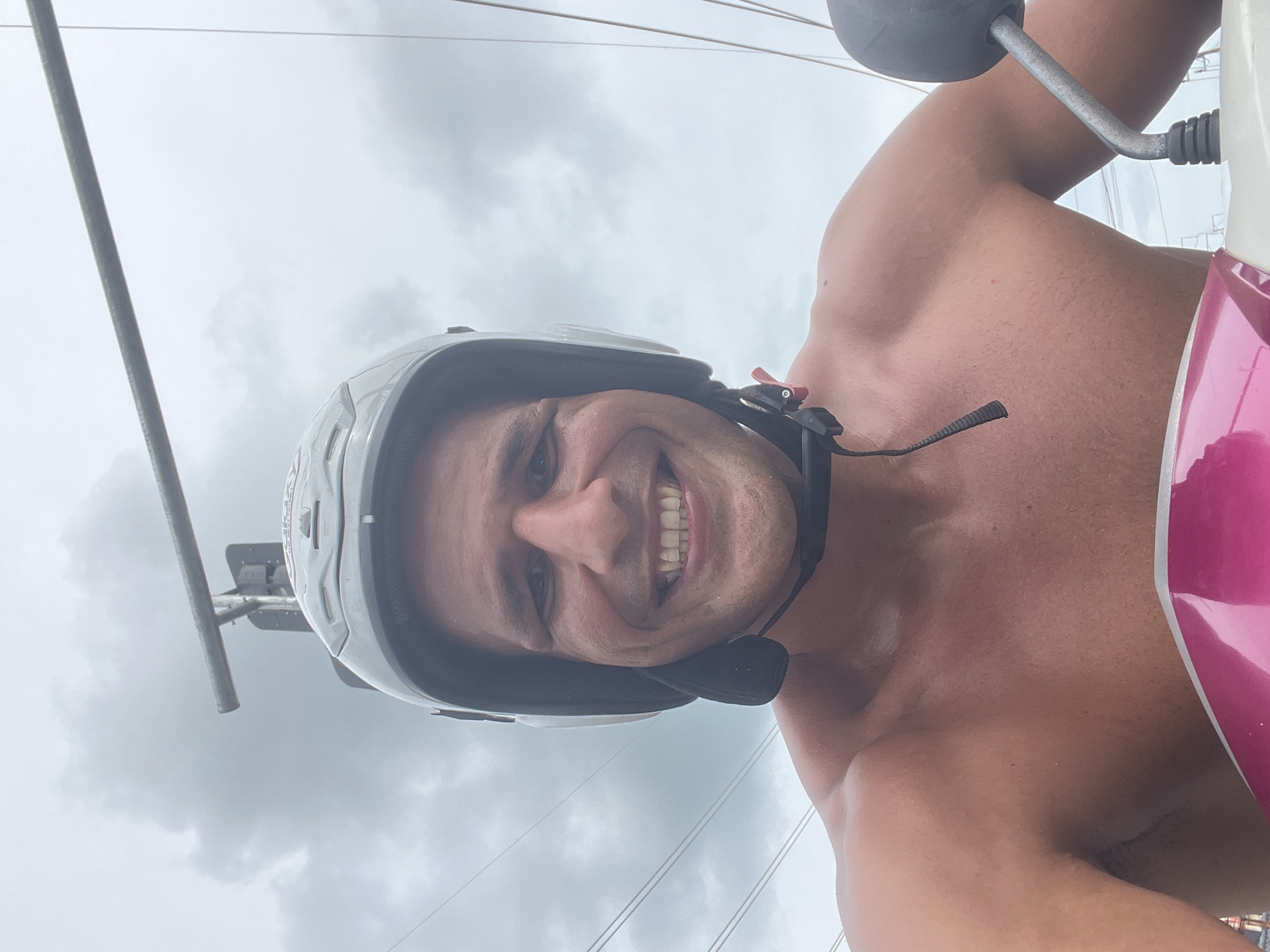 A shirtless man wearing a white helmet smiles while riding a scooter on a cloudy day in Chalong, Thailand. The man's chest and shoulders are visible, and he is looking directly at the camera. The scooter's pink and white body is in the foreground, and a metal handlebar is to the right. In the background, power lines and a traffic signal are visible against the overcast sky.
