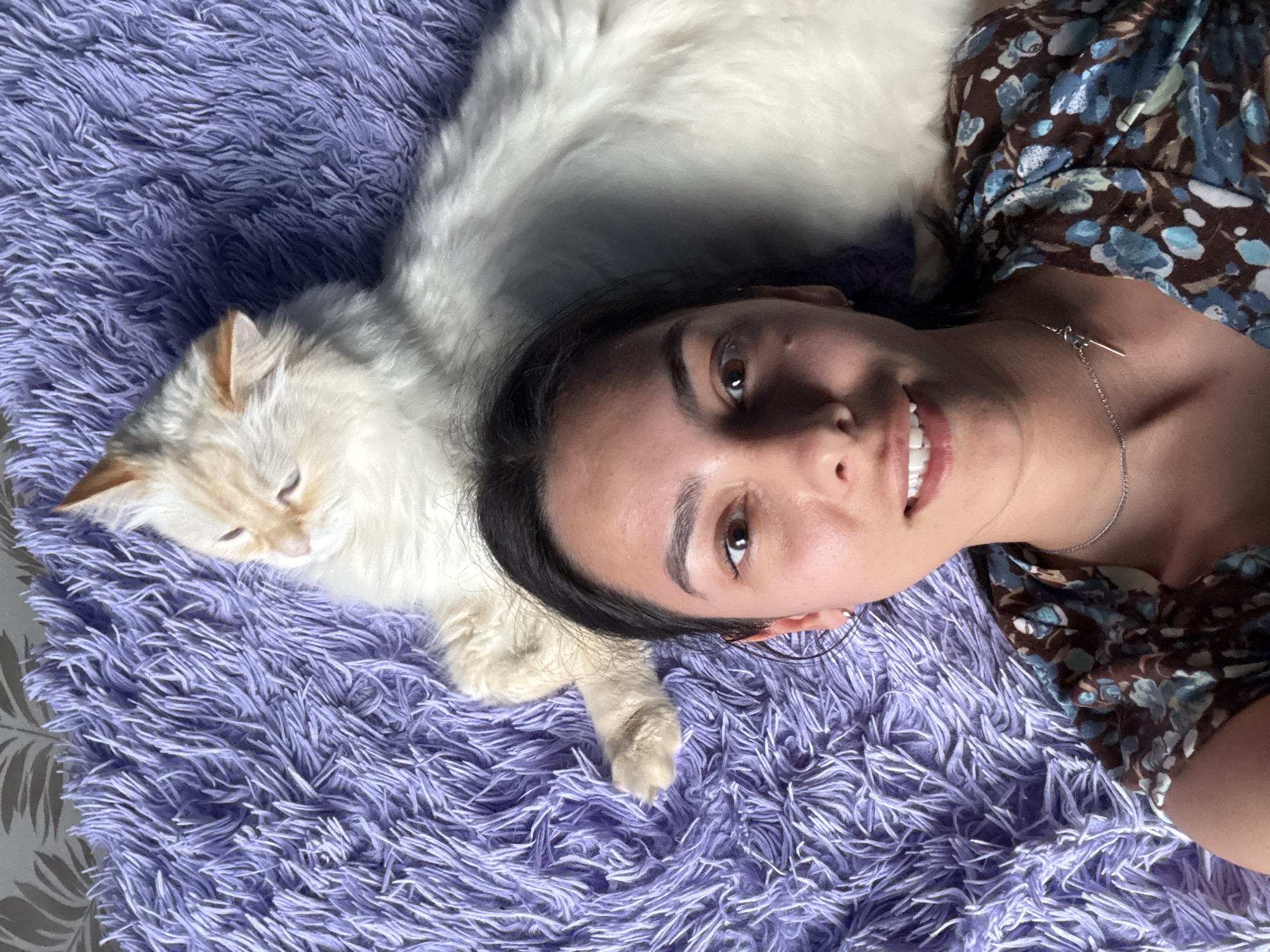 A selfie photograph shows a woman lying on a purple, shaggy rug with a light-colored cat. The woman has dark hair and is wearing a floral print dress and a silver necklace. She is smiling and looking at the camera. The cat is lying next to her, seemingly relaxed. The rug has a deep pile texture. The exact location in Chaikovsky, Russia, cannot be determined from the image alone.