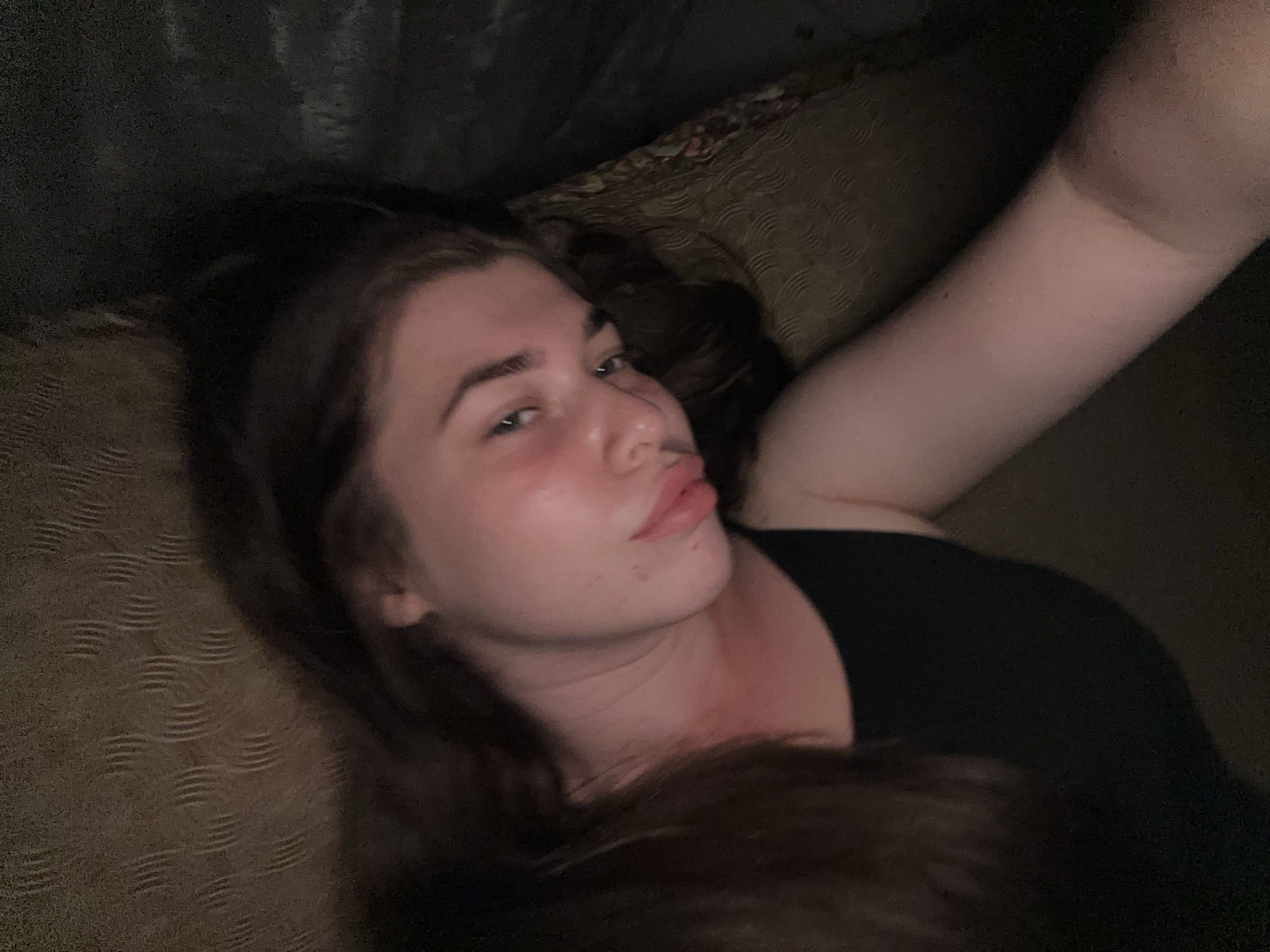 A young Caucasian female with long, dark brown hair is lying down, taking a selfie. Her head is tilted to the side, and her lips are pursed. She is wearing a black top. The background appears to be a dimly lit room with textured upholstery, possibly a sofa or bed. The lighting is low, casting shadows and creating a somewhat grainy effect on the image. The overall impression is of a casual, intimate moment captured by the subject herself. The location is unknown, and there is no information to suggest it is in Мещёры, Russia.