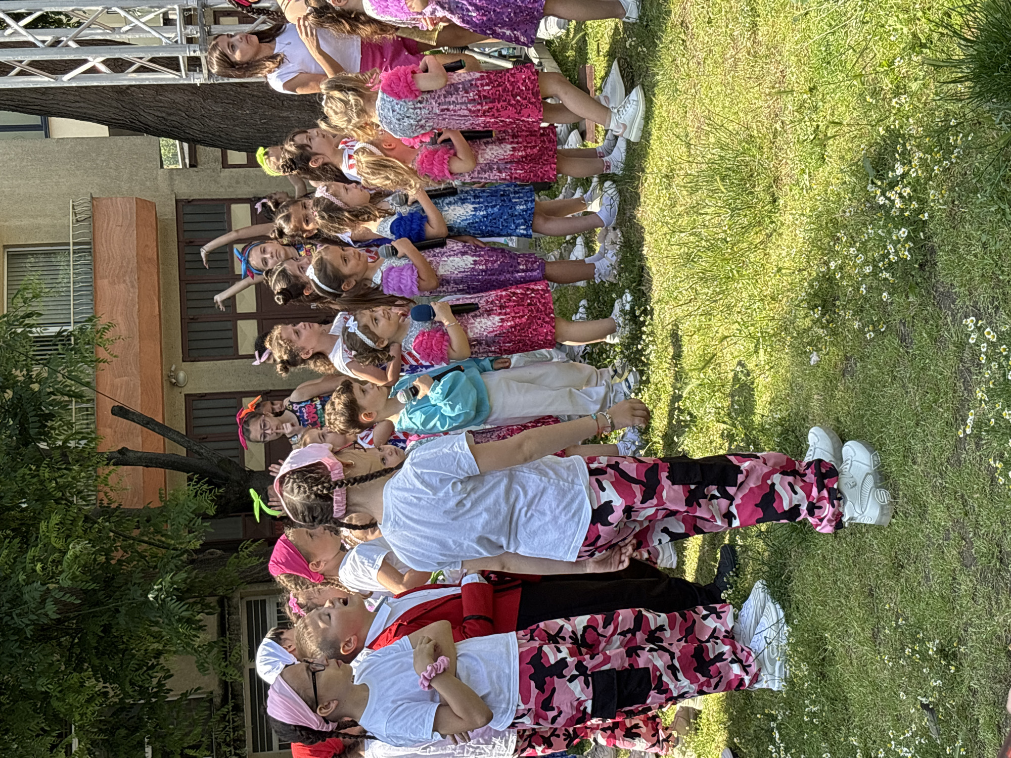 An outdoor scene in Varna, Bulgaria, depicts approximately two dozen children, predominantly girls, gathered on a sunny day. The ground is covered in green grass interspersed with small white flowers. In the foreground and left midground, several children, including a girl wearing glasses, are dressed in white t-shirts, pink and black camouflage cargo pants, and white athletic shoes; some also wear pink headbands. A boy in the left midground stands out in a red blazer, white shirt, and black pants. To the right midground, a line of five girls, each holding a microphone, wear sequined sleeveless dresses in varying shades of pink, purple, and blue. In the background, buildings with balconies are visible, alongside large green trees providing shade. On the far right, a silver metal stage truss structure is partially visible. A girl in the upper left background, wearing glasses, is making a "shhh" gesture with her finger to her lips. The children appear to be facing towards the right side of the frame, suggesting a performance or rehearsal. The specific location within Varna is not discernible from the visual information.