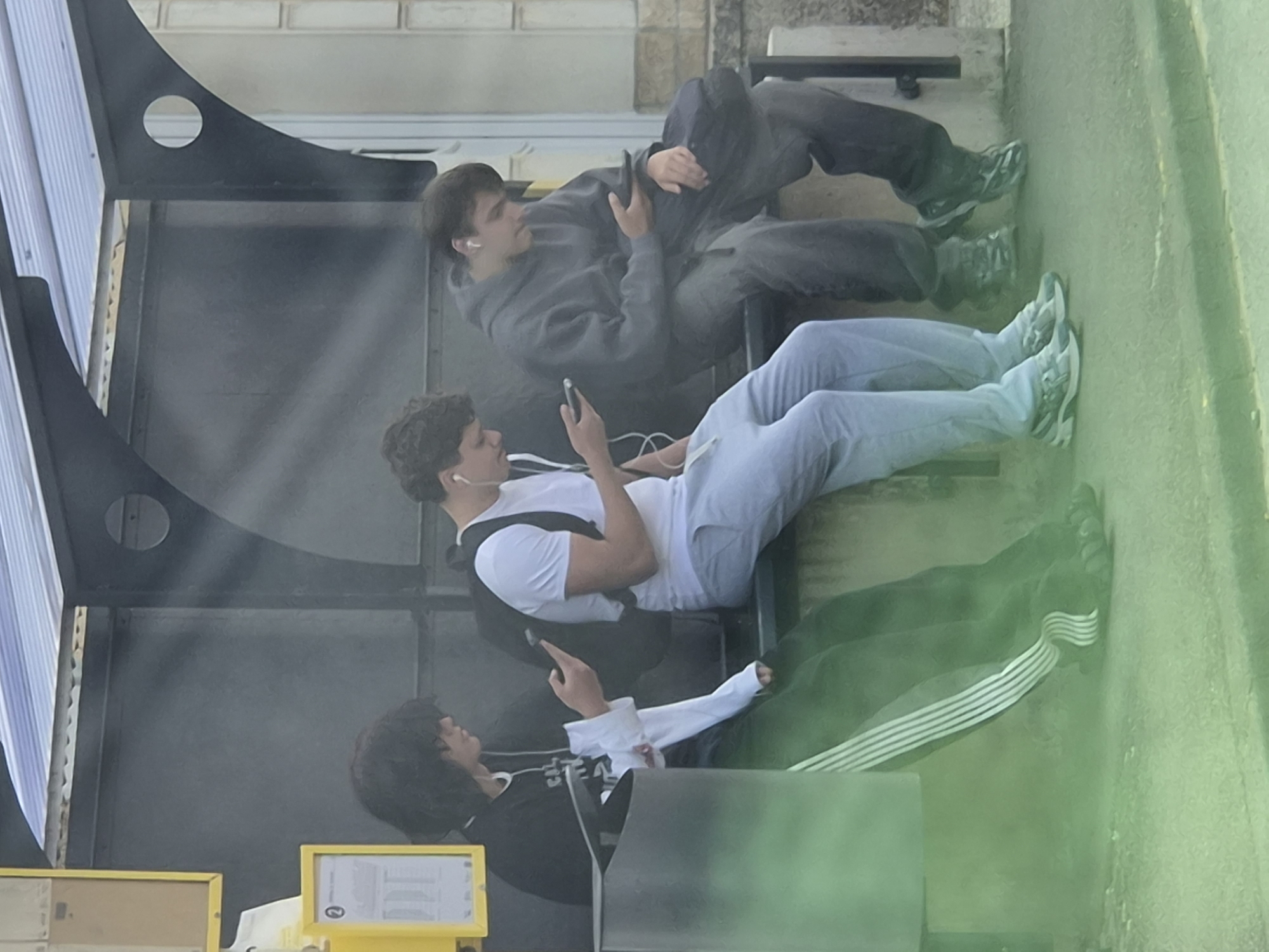Three individuals are seated side-by-side on a bench, likely at a bus stop, in Sanem, Luxembourg. The person on the left, with dark, shoulder-length hair, wears a black long-sleeved shirt with a white graphic and green pants with white stripes down the side. They are looking at their phone, holding it in their left hand. A white sleeve from a garment underneath is visible.
The middle individual has short dark hair and wears a white t-shirt and light blue sweatpants. They are looking down at their phone, which they hold in their right hand. A backpack is visible behind them.
The person on the right, with short brown hair, is wearing a grey hooded sweatshirt and dark pants. They are looking at their phone, which they hold in their right hand, and have white earbuds in. A dark bag rests on their lap.
The background appears to be a bus stop shelter with a metal roof and dark grey paneling. A yellow sign with a schedule is mounted on the wall to the left, and a circular detail is visible on the back panel of the shelter. The ground in front of the bench is a light grey surface, with a yellow line marking an edge, and is covered by a noticeable green haze, possibly due to a filter or lighting condition.