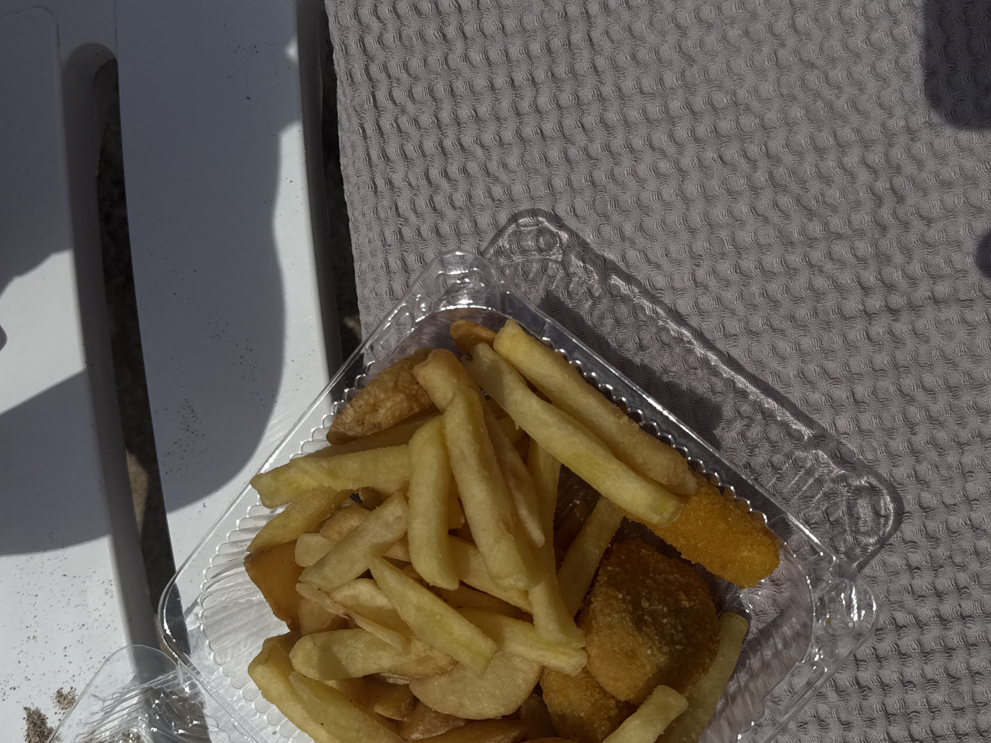 The image depicts a clear plastic container filled with fried food, specifically French fries and what appears to be fried fish pieces. The container rests on a textured gray surface, likely a fabric or towel. A portion of a white slatted object, possibly a beach chair, is visible on the left side of the frame. The scene is outdoors with harsh sunlight. Given the prompt's mention of Yevpatoriya, Ukraine, it's possible the image was taken in a park, beach or another outdoor area near that city. There are no people visible. The focus is on the food container.