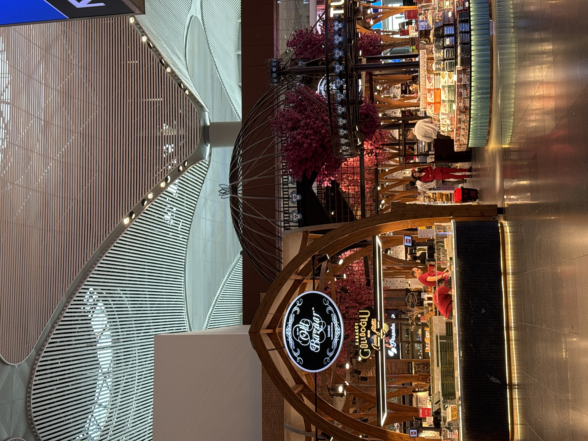 The image depicts an indoor marketplace, likely an airport or large retail space, in Arnavutköy, Turkey. The architecture features a modern, curved ceiling with white slats and prominent lighting fixtures. In the foreground, a stall with a wooden frame displays various packaged sweets and confections. A sign above the stall reads "Old Bazaar" with "Istanbul" and "Traditional Turkish Delicacies" below. Two individuals in red shirts are seen behind the counter, appearing to serve customers. To the right, another display area showcases more packaged goods, with several shoppers browsing. A woman in a red suit stands near a trash receptacle, while a man in a light-colored shirt stands behind a counter. The floor is a polished dark gray surface reflecting the overhead lights and the displays. The overall atmosphere suggests a bustling commercial environment with a focus on Turkish sweets and souvenirs.