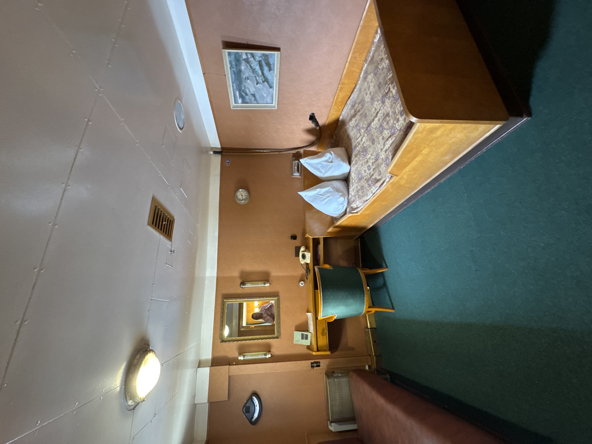 The image depicts a dimly lit room, likely a cabin, with a bed on the right and a desk with a chair on the left. The walls are painted an orange-brown color, and the ceiling is white with visible seams and rivets, suggesting an older structure or vessel. A light fixture is visible on the ceiling. The floor is covered with a dark green carpet.
On the right side of the room, a wooden bed frame holds a mattress with a patterned cover and two white pillows. A painting of a seascape hangs on the wall above the bed. A coiled hose-like object extends from the wall near the painting towards the bed.
On the left side, a wooden desk is positioned against the wall. It has a drawer and a telephone with a rotary dial. A green upholstered chair with a wooden frame is placed in front of the desk. A mirror hangs above the desk, reflecting a person holding a phone. To the left of the desk, a radiator or heating unit is visible against the wall. A black, wall-mounted object, possibly a lamp or fixture, is also present on the left wall.
A narrow, reddish-brown upholstered bench or sofa is partially visible on the far left of the image.
The overall impression is of a compact, functional living space, potentially on a ship or in a historic building. No specific location in Saint Petersburg can be identified from the visual cues within the image.