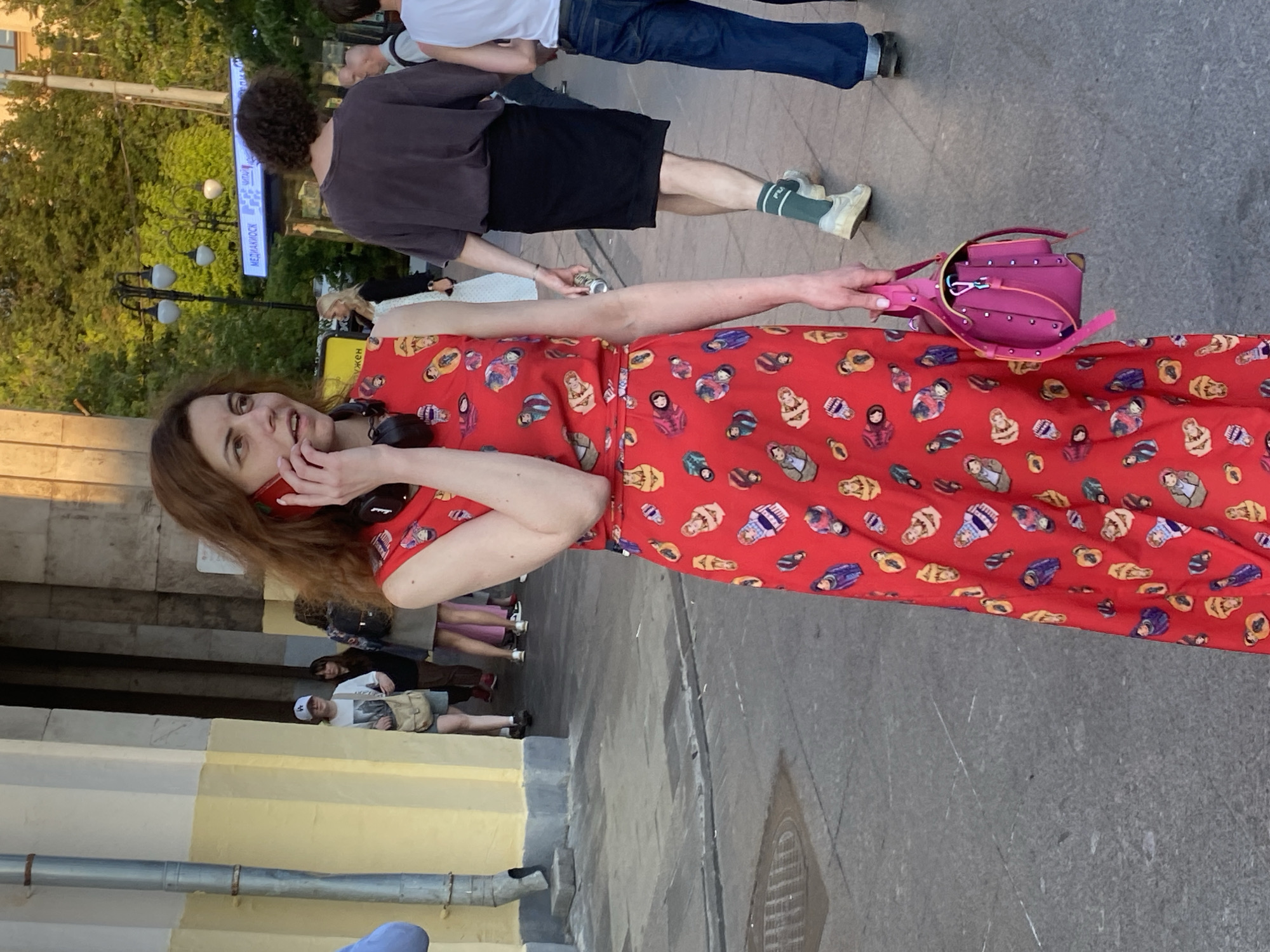 A woman with reddish-brown hair is standing outdoors in a city and talking on a red mobile phone. She is wearing a sleeveless red dress with a pattern of matryoshka dolls. She has black headphones around her neck and is holding a bright pink handbag. In the background, other people are walking, and there is a sign with Cyrillic text that reads "МЕДИАМОСК" (MEDIAMOSK). This indicates the location is Moscow, Russia, likely near a metro station entrance as the sign appears to be for a media kiosk. The environment suggests a public urban space with paved ground and a building with yellow and gray architectural elements. The lighting indicates it is daytime, possibly late afternoon or early evening due to the warm light.