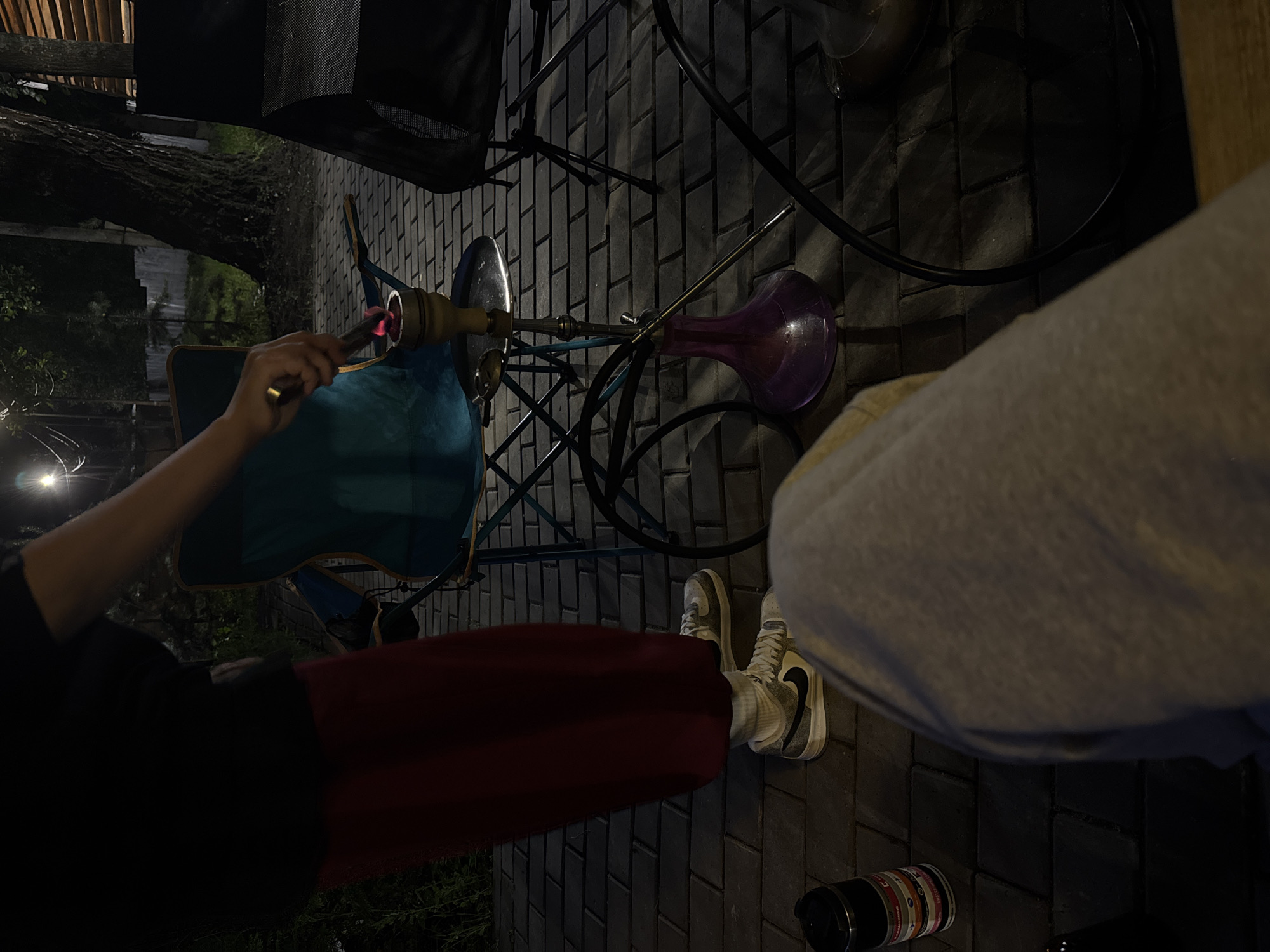 An overhead shot captures a person from the waist down, with their left leg extended and right leg bent. They are wearing a dark t-shirt and a dark red skirt, white socks, and white sneakers with black accents. The person's hand, holding a metal utensil, is positioned over a hookah. The hookah features a purple base and a metallic stem, with a charcoal-filled bowl at the top. A teal folding chair is partially visible to the left of the hookah, and another dark chair is to the right. The scene is set on a paved patio made of dark grey bricks. In the background, a large tree with rough bark stands prominently, its branches extending upwards. The lighting suggests it is nighttime, with some ambient light illuminating the scene. A black thermal mug with colorful bands is placed on the ground to the left of the frame. The overall atmosphere is relaxed and informal. The location is Doma 1096 Km, Russia.