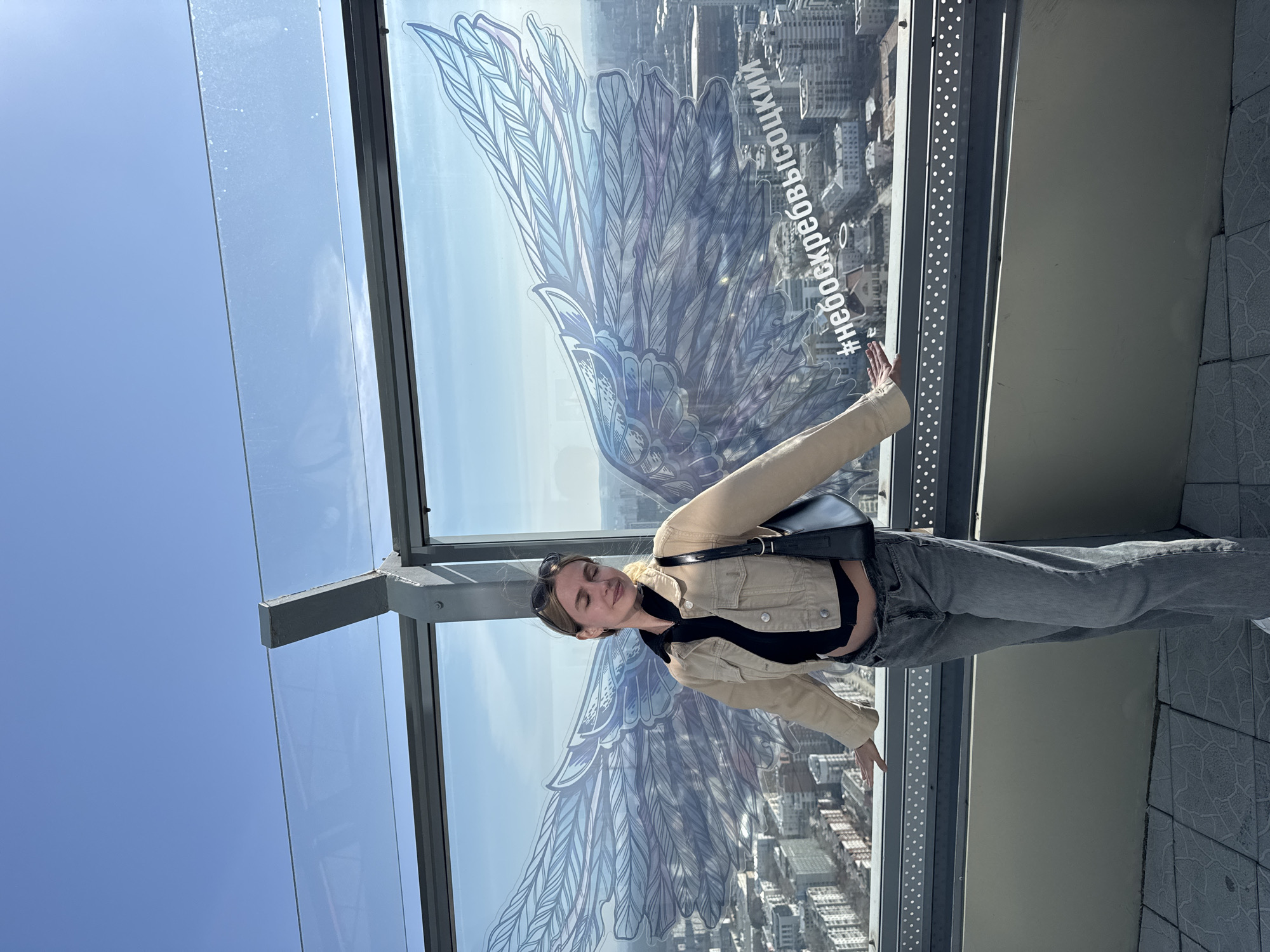 A full-body shot presents a fair-skinned woman in casual attire—a light beige jacket, a black top, grey jeans, and a black shoulder bag. She poses against a glass window on which a large wing design is drawn. The window frames a high-altitude view of a cityscape. A text inscription, possibly a hashtag, is visible on the right side of the window. The sky is a clear blue and the image is taken during daylight.