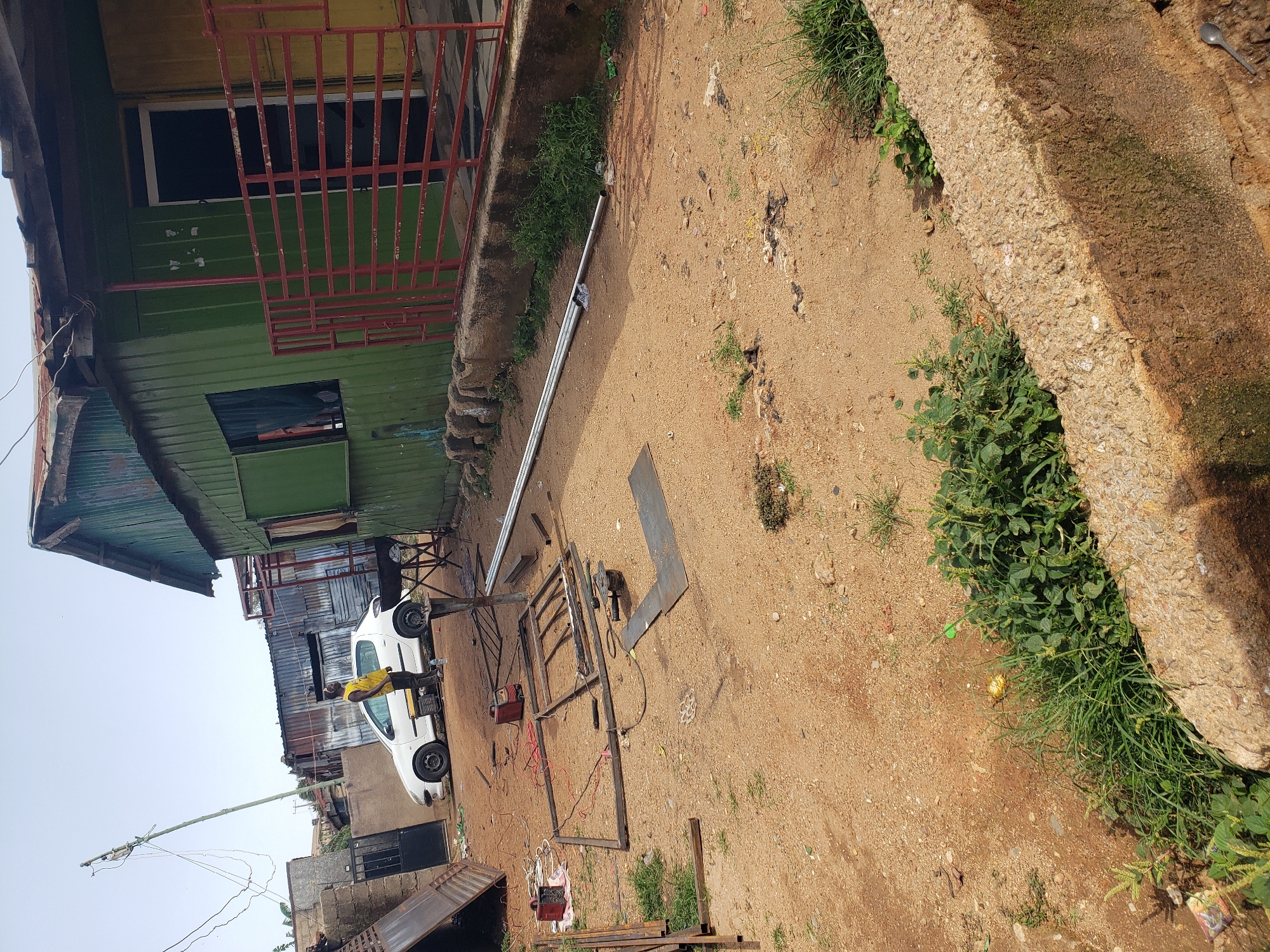 This is an outdoor scene on a sandy lot, possibly in the city of Jos, Nigeria, with a green corrugated metal building and a white car. A man in a yellow tank top and dark pants stands near the car, looking towards the left of the frame. In the foreground, various metal pieces, including a welding machine, are scattered on the ground, suggesting construction or repair work is underway. A metal frame is partially assembled. The building has a red-railed porch and an open doorway. Vegetation, including grass and some larger plants, is present in patches around the scene, and a concrete barrier runs along the lower edge of the frame. The sky is clear and bright.
