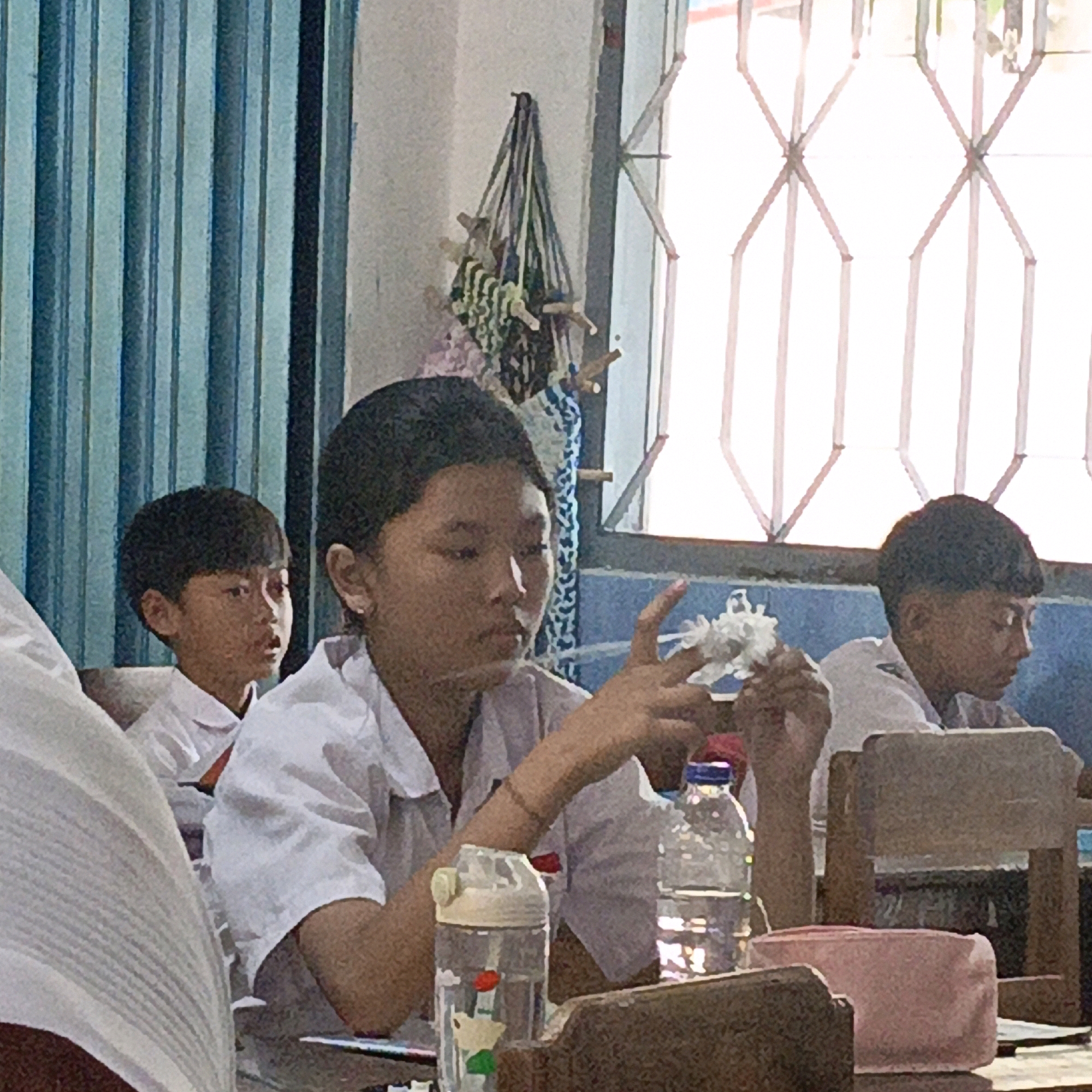 In Salatiga, Indonesia, inside what appears to be a classroom setting, three young students are observed. The central figure, a girl wearing a white school uniform, is facing left and looking down, with her right hand raised, fingers splayed as if in thought or gesturing. Beside her, on the right, a boy in a similar uniform is also looking down. To the left, partially obscured, another boy in a white uniform is visible, looking towards the right. On a table in the foreground, several objects are present: a clear water bottle with a red lid and a cartoon character inside, another clear water bottle, and a pink rectangular object, possibly a pencil case. The background features blue striped curtains on the left and a window with a diamond-patterned metal grate on the right, allowing bright light to enter. A decorative hanging item with numerous small, colorful elements is suspended on the wall to the right of the curtains.