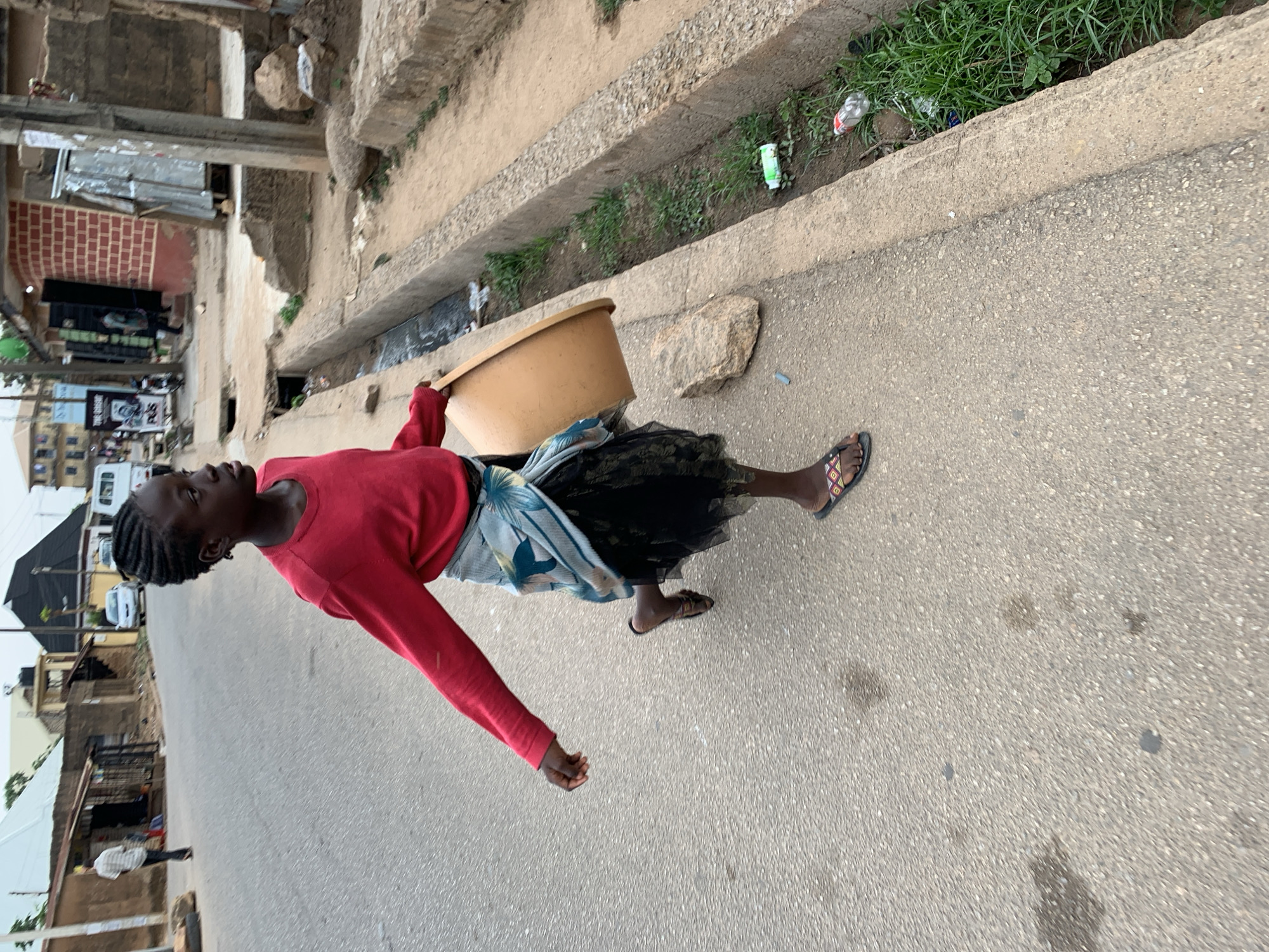 The image depicts a daytime street scene, likely in an urban or semi-urban area of Jos, Nigeria. The central subject is a young person, appearing to be female, with dark skin and braided hair, walking on a paved road. She wears a long-sleeved red top, a patterned light blue and white wrap-around cloth over a black skirt, and open-toed sandals with a colorful geometric pattern on the strap. She carries a large, light brown plastic basin or bowl tucked under her left arm.
To the right of the road, there is a concrete curb separating the road from a wide gutter or drainage ditch. This ditch contains green weeds, some litter including plastic bottles, and a few rocks.
In the background, the paved street continues, lined by various buildings. These structures show diverse architecture, including brick facades, concrete walls, and some smaller, possibly temporary, shacks with corrugated metal elements. A white van or bus is parked further down the street, and other indistinct figures of people can be seen in the distance, suggesting a bustling environment. The lighting indicates natural daylight, with no harsh shadows, suggesting a clear day. No prominent or legible text is visible in the frame.