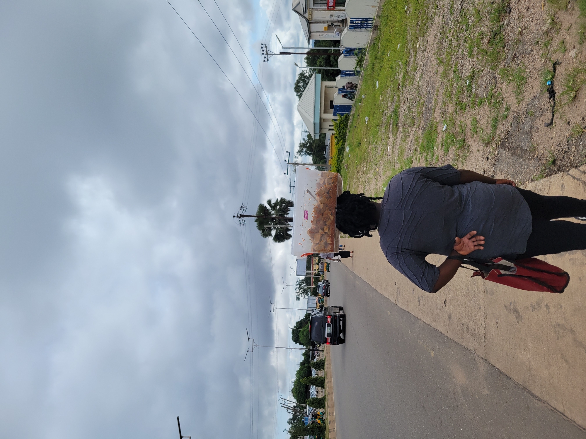 A woman walks along a sidewalk in Jos, Nigeria, carrying a large clear plastic container filled with food items on her head. She is wearing a dark grey shirt and black pants, with a red bag slung over her left shoulder. The street is paved and lined with trees and buildings. Several vehicles, including cars and a tricycle, are visible on the road. The sky is overcast with clouds. The scene is one of everyday street life in a Nigerian city.