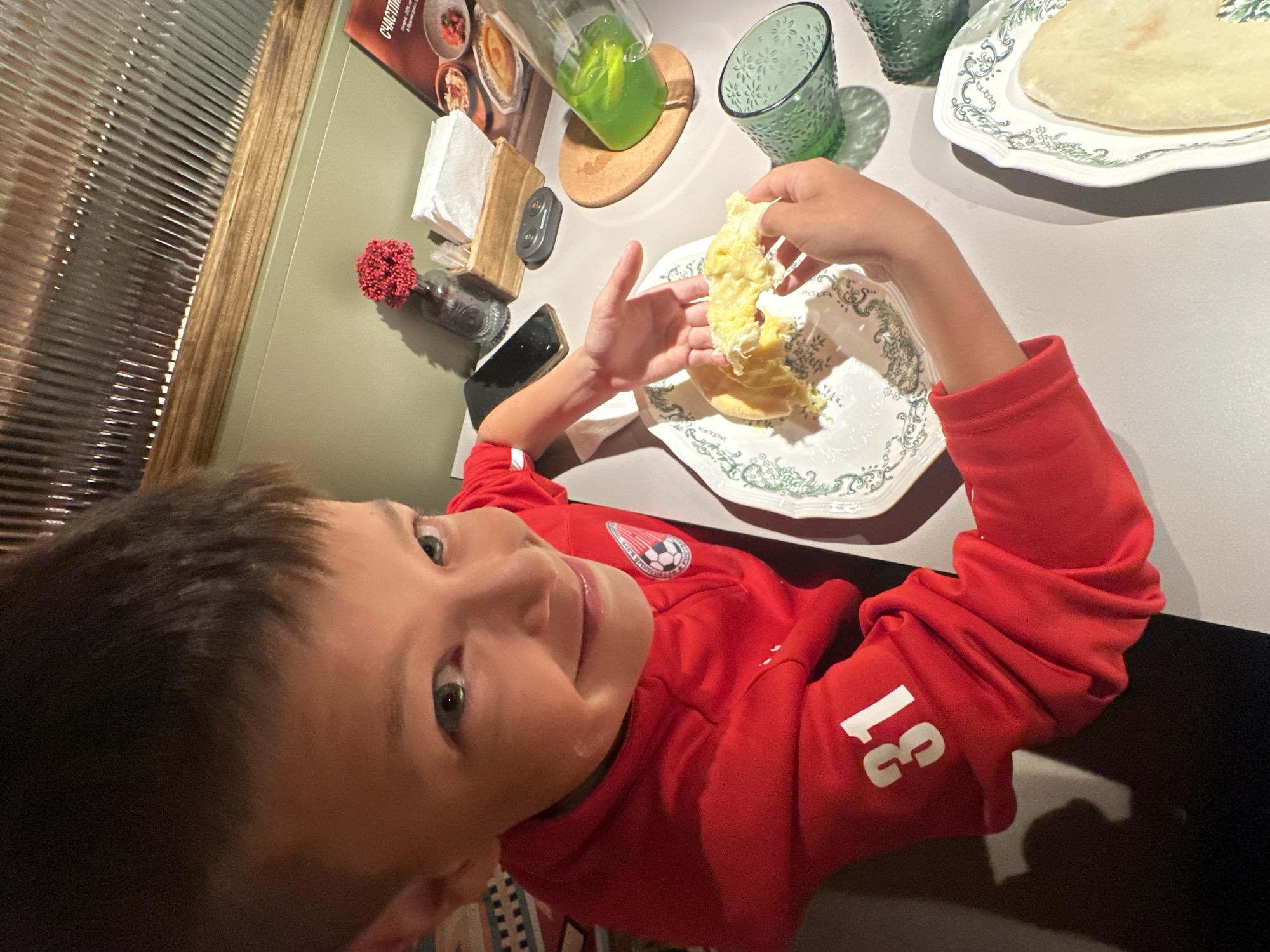 The image depicts a young boy, estimated to be between 5 and 10 years old, seated at a light-colored table in a restaurant or cafe located in Chaikovsky, Russia. He has short brown hair, green eyes, and is smiling directly at the camera. He wears a red, long-sleeved athletic shirt featuring a partial zipper at the neck. A circular logo with a soccer ball design is present on the left chest, and the white number "31" is printed on the right sleeve. The boy is actively holding and consuming a portion of a yellow, fluffy dish, consistent with scrambled eggs or an omelette, using both hands.
On the table in front of him, two white plates with green decorative patterns are visible. One plate holds the food the boy is eating, while another, partially visible on the right, contains a flatbread or tortilla. A clear pitcher filled with bright green liquid rests on a round wooden coaster, alongside two empty green textured glasses. Additional items on the table include a black smartphone placed face down, a small wooden container holding white paper napkins, a black electronic device (likely a service pager), and a small glass vase with red decorative elements. A menu or promotional leaflet, partially visible on the far right, displays food images and the Cyrillic word "СЧАСТИЕ" (Happiness).
The background consists of a two-toned wall, with a lighter green lower section separated by a horizontal wooden trim from a darker green upper section. Above this, a reflective, vertically fluted or corrugated panel, potentially a window or decorative wall covering, is present. A patterned fabric or rug is partially visible on the lower left side of the frame.