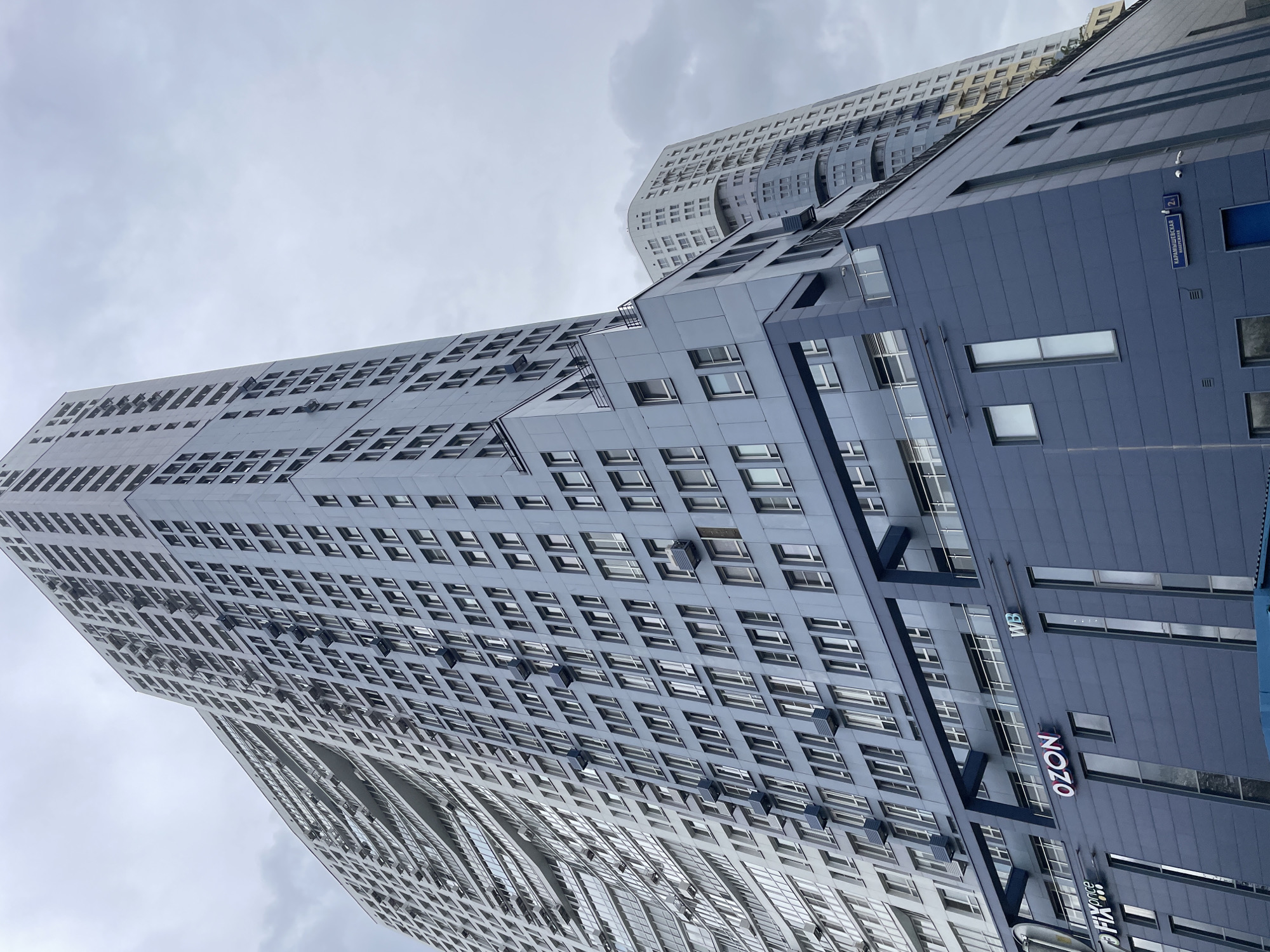 A low-angle shot reveals two modern, high-rise buildings in Khimki, Russia, set against a cloudy sky. The buildings are constructed with a geometric pattern of numerous square windows. The building on the right is colored dark blue and has storefronts displaying the logos "OZON", "WB," and "FIX Price." A blue street sign reads "КАРАМЫШЕВСКАЯ НАБЕРЕЖНАЯ 24". The building on the left is gray with a more complex and curvilinear design. Air conditioning units are visible beneath some of the windows. No people are visible in the frame.