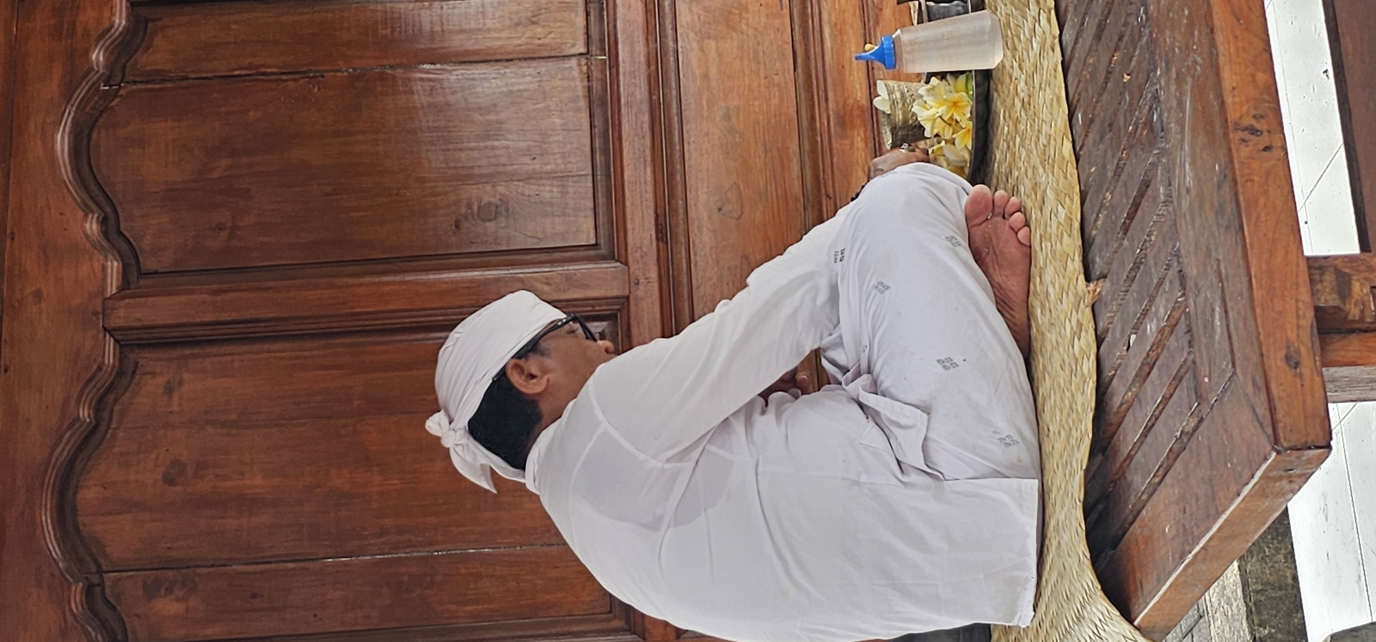 A person wearing white clothing and a white head covering is kneeling in what appears to be a place of worship. The location is identified as Kuta Selatan, Indonesia. The individual is positioned in front of a wooden structure, likely a door or shrine, with offerings placed at the top. The offerings include a small bottle of water and flowers. The person's feet are visible on a woven mat.