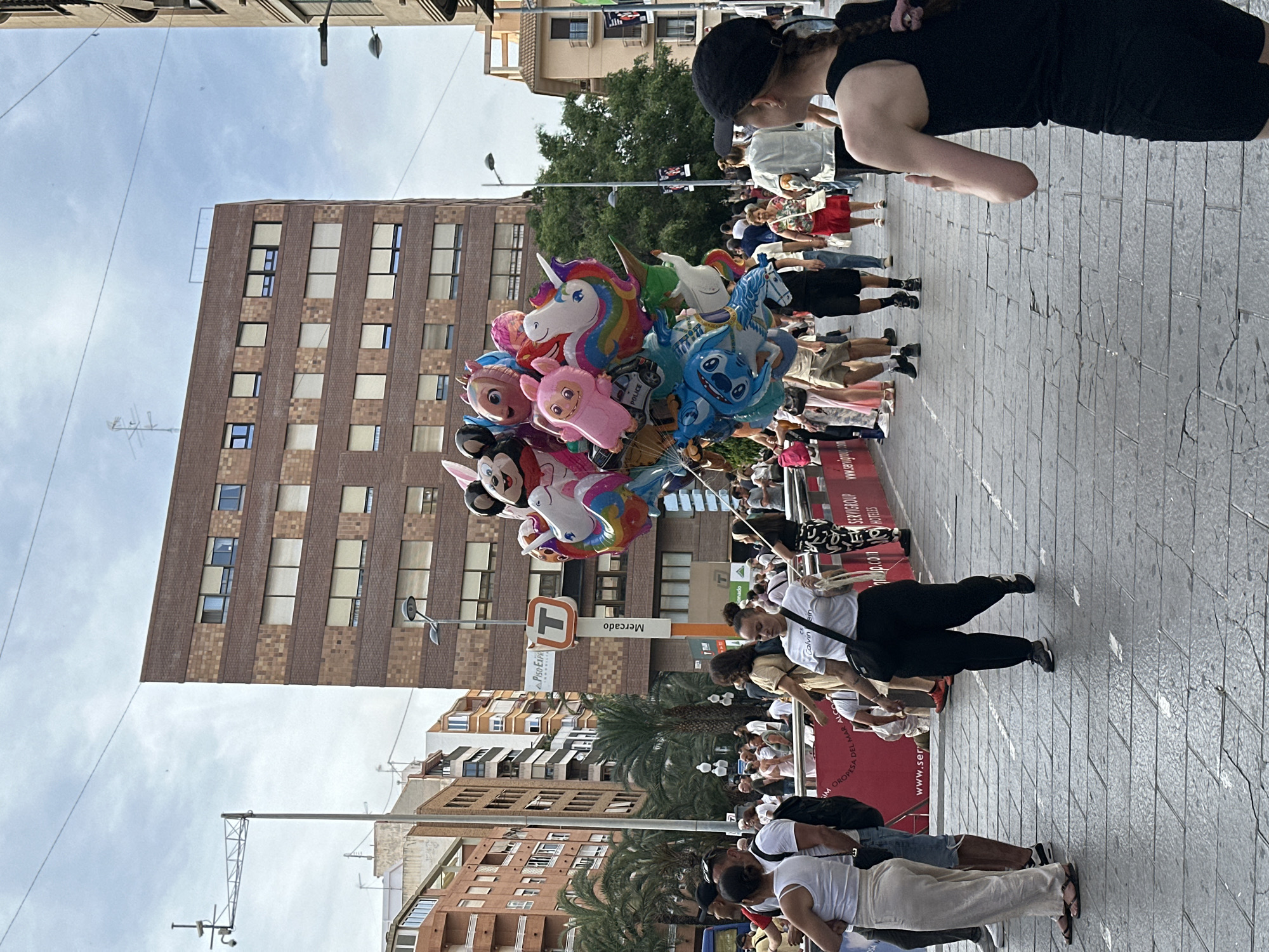 The image captures a bustling urban street scene at the Mercado tram stop in Alicante, Spain, under an overcast sky. A large, colorful cluster of foil balloons, featuring characters such as Mickey Mouse, multiple unicorns (one rainbow-colored), and Stitch, dominates the central mid-ground, carried by an individual largely obscured by the balloons. In the left foreground, a woman in a white tank top and light-colored trousers stands beside a man in a white t-shirt and denim shorts. A woman in a white "Calvin Klein" t-shirt and black pants walks towards the left, carrying a black shoulder bag. On the far right, a person wearing a black tank top and a black baseball cap, with braided hair, is seen from behind, looking leftward. Numerous pedestrians in casual attire are dispersed across the scene, walking in various directions on the cracked, rectangular stone pavement.
The background features several multi-story buildings. A prominent brown brick building with alternating horizontal bands of lighter bricks and multiple windows, many equipped with beige roller blinds, stands centrally. To its left, other buildings and several palm trees are visible. A white tram stop sign, bearing an orange "T" logo and the word "Mercado" vertically, is positioned in the mid-ground. Overhead utility wires traverse the sky. Red banners or barricades, partially displaying text like "www.ser" and "servigroup.com," are present in the lower mid-ground. A streetlight and a CCTV camera mounted on tall poles are also visible.