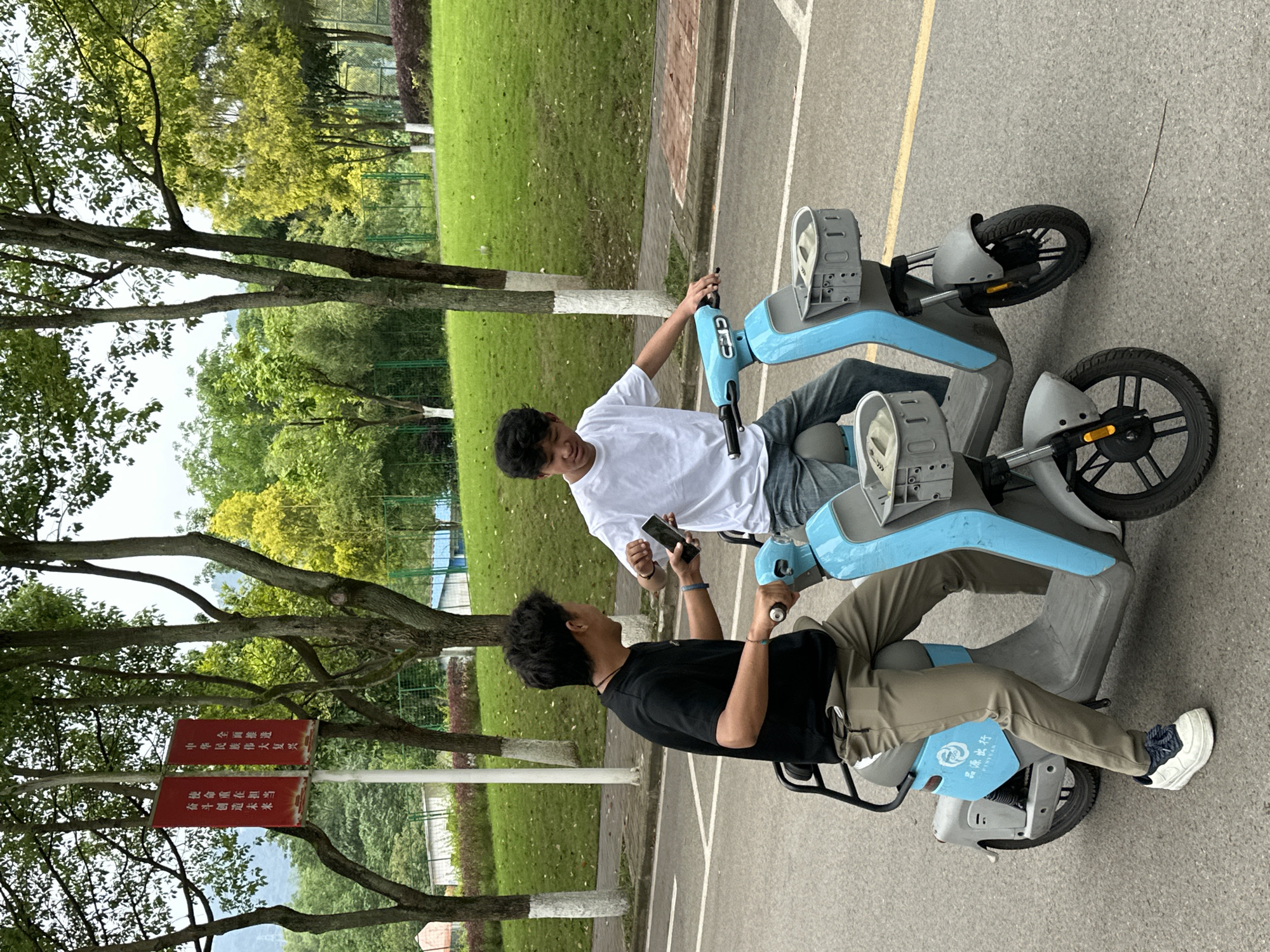 Two young men are depicted outdoors, seated on light blue and grey electric scooters on a paved path. The man on the left, wearing a white t-shirt and jeans, looks towards his companion. The man on the right, dressed in a black t-shirt and khaki pants, holds a smartphone and appears to be interacting with the first man. They seem to be pausing during or preparing for a ride.
The setting is a green and treelined area, likely a park or campus in Jiangjin, China, with lush grass and various trees. Distant mountains are faintly visible through the foliage. The paved path features painted white and yellow lines. The overall impression is a relaxed, daytime scene with comfortable weather.
Notable details include a red sign positioned among the trees on the left, displaying Chinese text. The text reads: "全面推进" (Comprehensively promote), "中华民族伟大复兴" (The great rejuvenation of the Chinese nation), "使命重在担当" (Mission lies in responsibility), and "奋斗创造未来" (Struggle creates the future). One of the electric scooters features a blue panel with the brand name "品源出行" (Pinyuan Travel/Pinyuan Ride) and "PINYUAN" in English, indicating a shared mobility service.