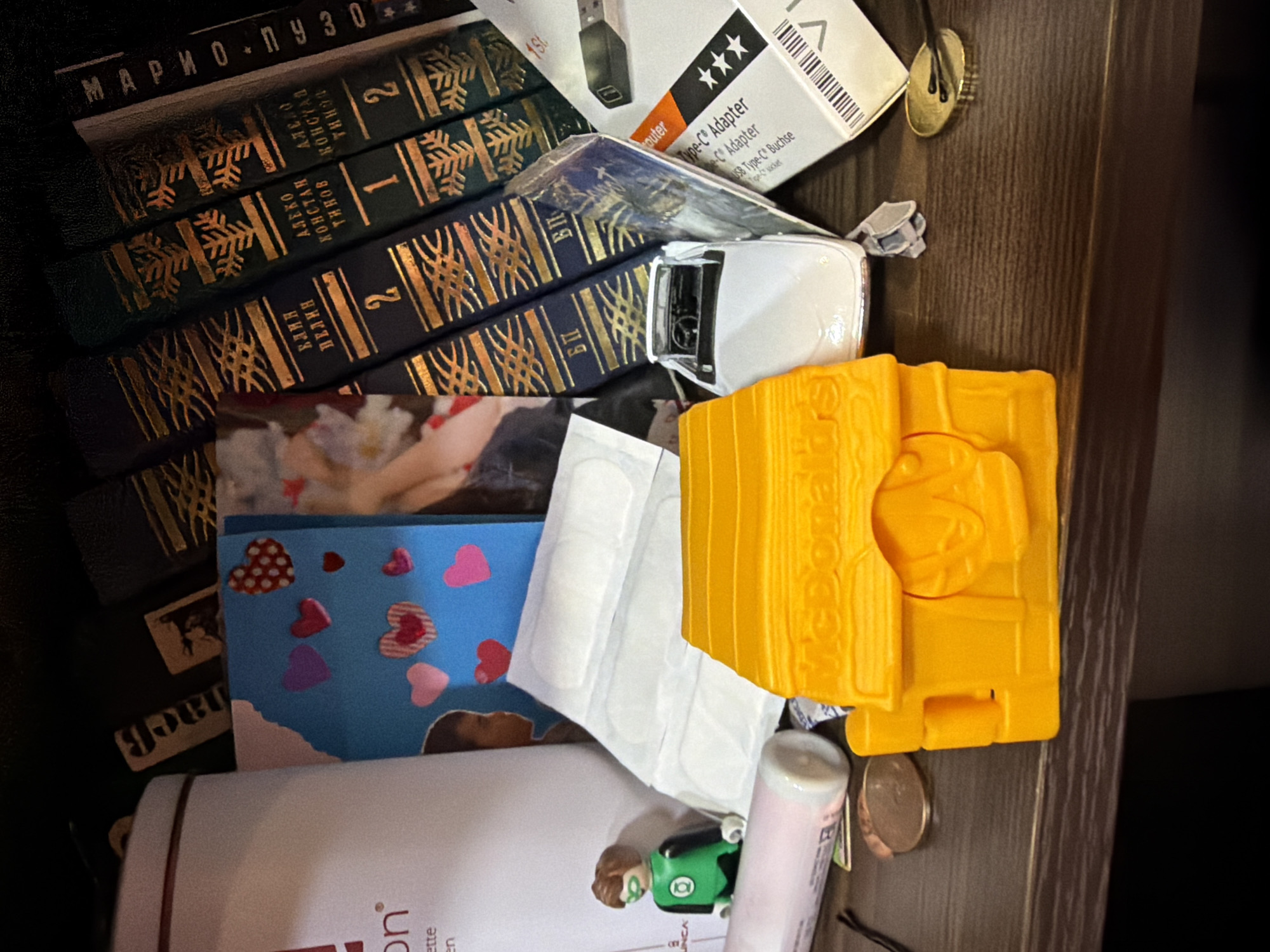 This is a cluttered scene containing various objects, some overlapping. There are several books, with the author "Марио Пузо" visible on the spine of one. The books are dark green with gold ornamental designs. A white box labeled "Type-C Adapter" is also present. A small white toy car and a bright yellow toy with the McDonald's logo are also visible. Additionally, there's a handmade card adorned with heart stickers. A white container with "on" visible on the label sits near a tube of lip balm and a small Green Lantern figurine. A few coins are scattered among the objects. The scene appears to be on a dark brown wooden surface.