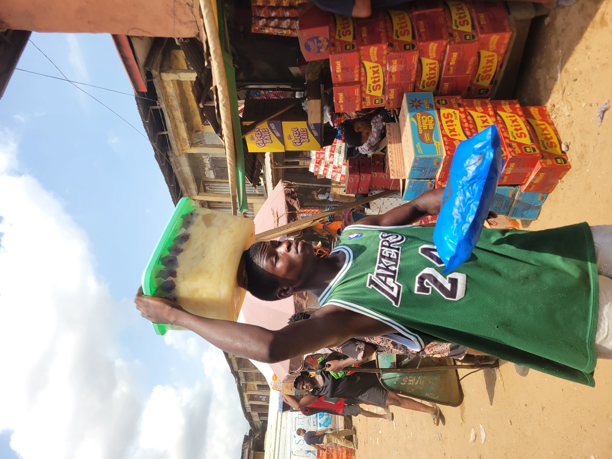 A street-level view in Onitsha, Nigeria, shows a man wearing a green basketball jersey with the word "LAKERS" and the number "24" standing in a busy commercial area. He balances a large, rectangular container with a green lid on his head. The container's contents appear to be a light-colored food item. He also holds a blue plastic bag in his left hand. Stacks of boxes, labeled "Stixi," and "Cheese Balls PARTY PACK" are displayed for sale behind him. Other pedestrians and a person pushing a wheelbarrow are visible in the background. The location appears to be a marketplace with buildings and stalls lining the street. The sky is partially visible with clouds.