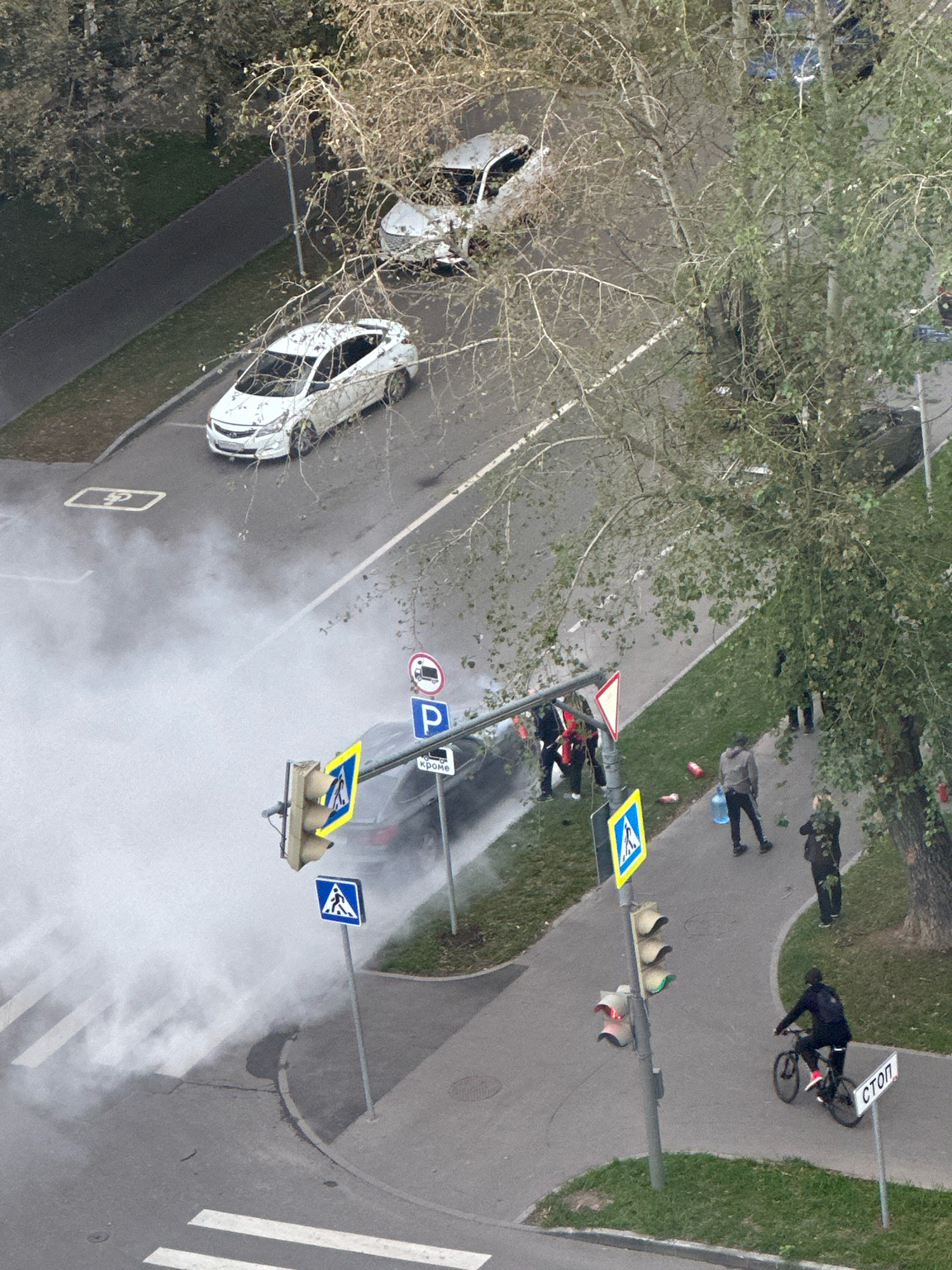 An aerial view captures a street scene in Moscow, Russia, with a black car appearing to be involved in an incident, emitting a significant amount of smoke. The smoke billows across the road and sidewalk, obscuring parts of a pedestrian crossing. Several individuals are present near the car and on the adjacent sidewalk, some observing the situation, one of whom is carrying a large water bottle. Traffic signals are visible, with one showing a red light. A cyclist is riding on the sidewalk, passing a "STOP" sign. Two white cars are parked on the street, one of which is partially obscured by trees. The environment includes trees with sparse foliage, suggesting an autumn or early spring setting, and a paved sidewalk and road.