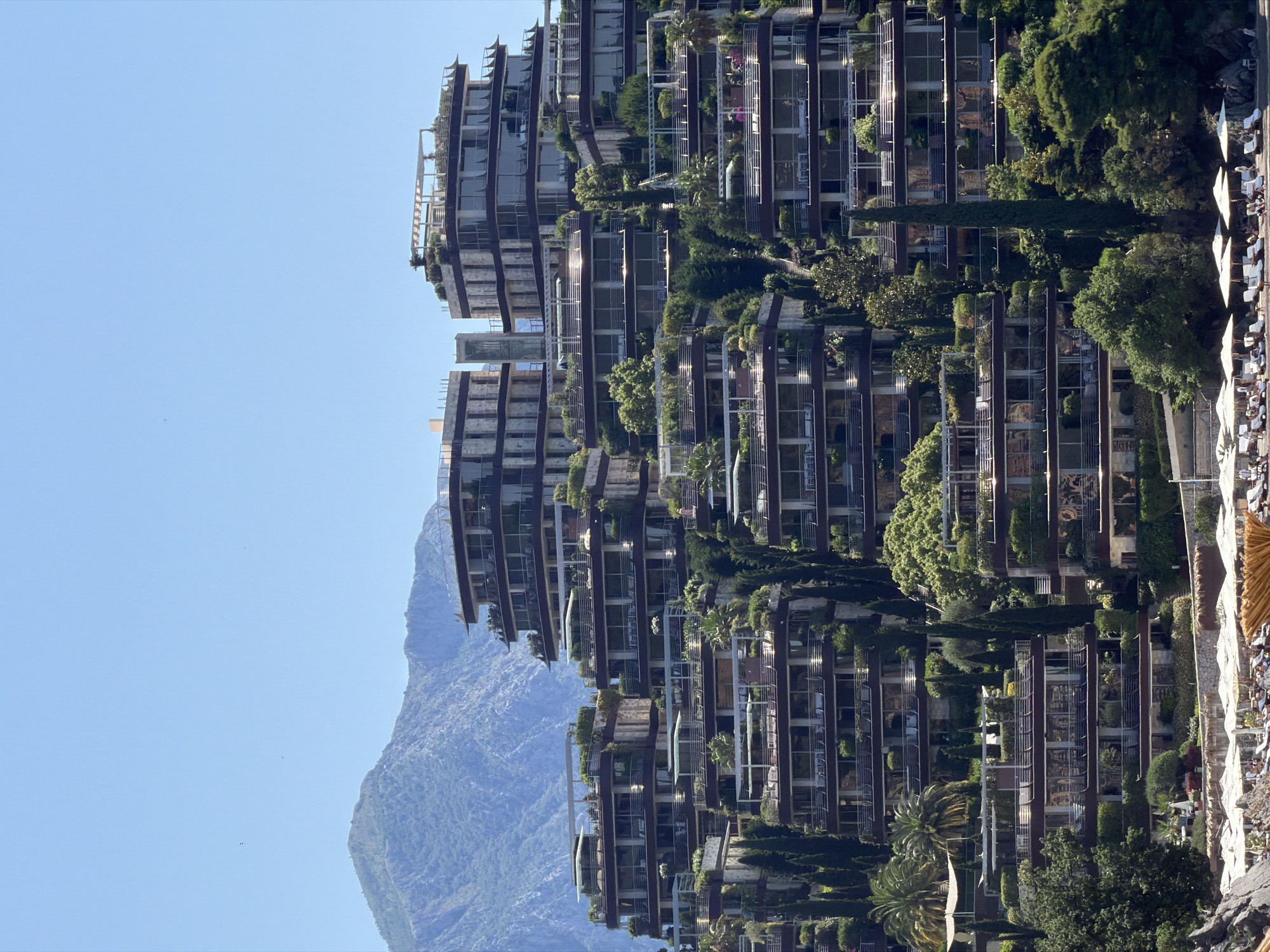The image displays a large, modern, terraced residential complex, identified as Dukley Gardens, situated on a verdant hillside in Budva, Montenegro. The complex consists of multiple multi-story buildings cascading down towards a beach.
Each building features extensive glass windows and wide, deep balconies or terraces, almost all of which are heavily landscaped with lush greenery, including trees, shrubs, and flowering plants, creating a "hanging gardens" effect. Tall, slender cypress trees are interspersed throughout the complex, providing vertical elements. Rooftops of some buildings include pergolas or shaded structures.
In the background, a substantial mountain range with rugged, vegetated slopes rises under a clear, bright blue sky. Several small, dark specks, possibly birds, are visible against the sky.
In the foreground, at the base of the hillside, a beach area is visible. Numerous white beach umbrellas are neatly arranged in rows, accompanied by white lounge chairs. Scattered individuals are present on the beach, engaged in relaxation. A dark rock formation borders the left side of the beach. The sunlight is bright, casting clear shadows, indicating midday or early afternoon.