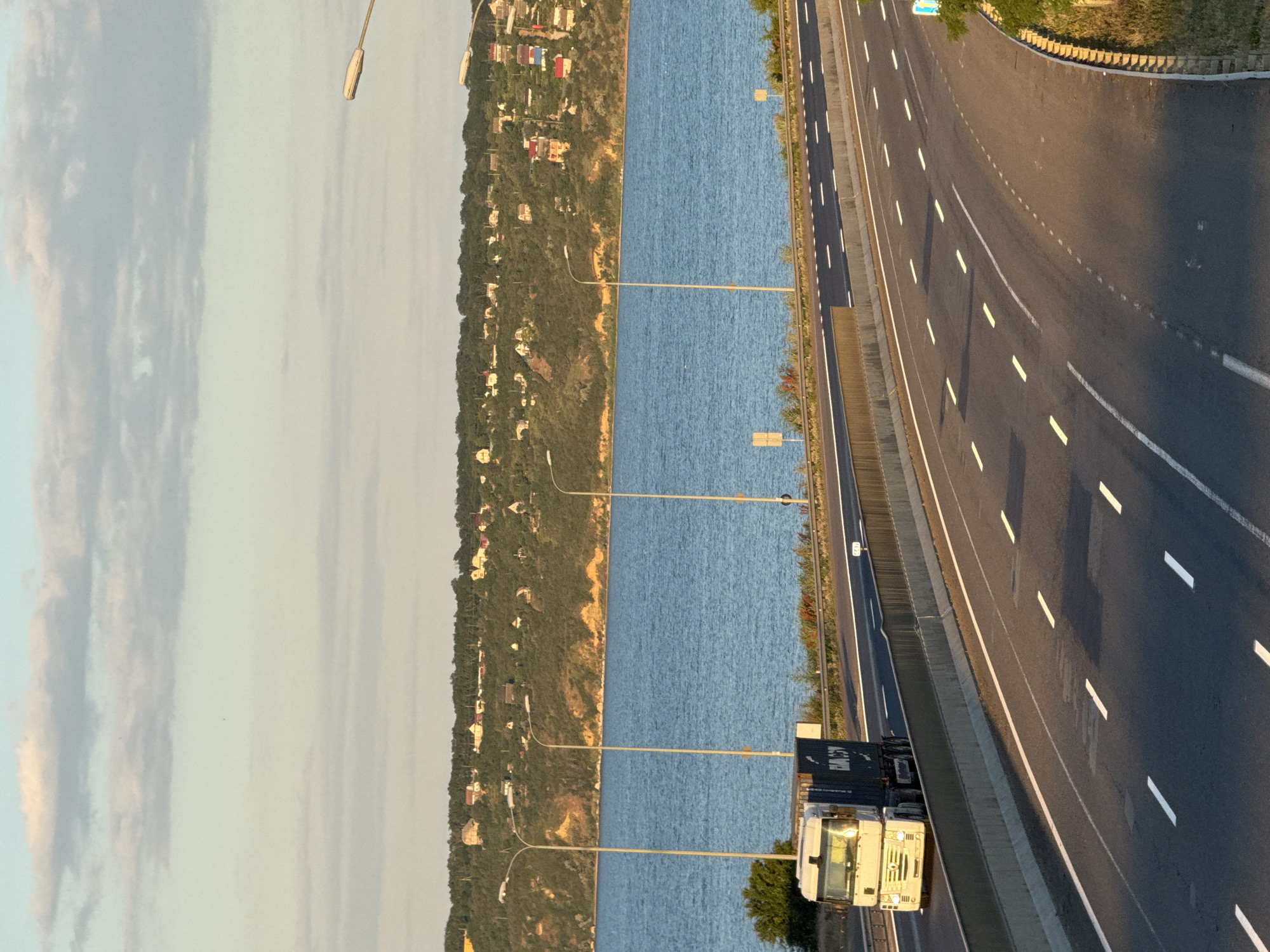 A wide-angle, eye-level shot captures a semi-trailer truck traveling on a multi-lane highway. The truck, a white cab with a dark blue container, is in the left lane. The highway curves to the right, with white dashed lines marking the lanes. On the left side of the highway is a steep, tree-covered hill dotted with houses. The hill overlooks a wide, calm body of blue water, possibly a river or lake, with more houses visible on the distant shore. The sky above is a pale blue with scattered, wispy clouds. Streetlights are visible along the highway and on the hill. The scene appears to be set in Altestove, Ukraine, with the lighting suggesting it is late afternoon or early evening.