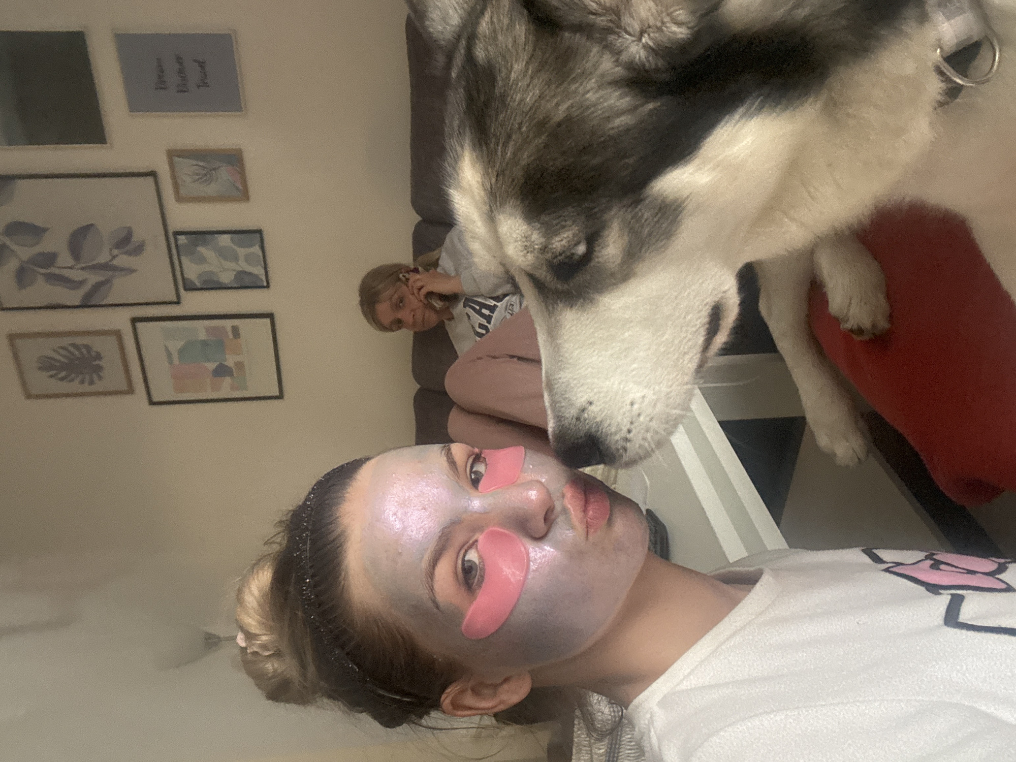 An indoor scene in Plovdiv, Bulgaria, depicts two individuals and a dog in a relaxed home environment. In the foreground, a young person with light hair pulled back wears a shimmering purple face mask and pink, gel under-eye patches. They look calmly towards the viewer. A black and white husky-type dog, prominently featured in the upper right, is very close to this person's face, appearing to sniff or observe them with interest.
In the mid-ground, another person with blonde hair is reclining on a dark sofa, holding a phone to their ear as if engaged in a call. Part of their light-colored shirt is visible, displaying the letters "GA".
The background features a light-colored wall decorated with several framed pictures. One notable frame is blue with white text reading "Dream Gather Travel." Other frames contain various artworks, including botanical designs with light blue leaves and a tropical leaf, as well as abstract patterns and shapes. A red cushion is visible under the dog's body, and a white furniture piece is partially seen. The overall mood is casual and domestic.
