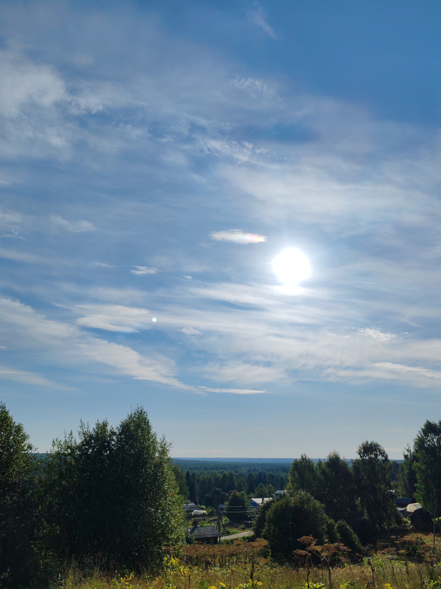 The image depicts a rural landscape under a partly cloudy blue sky. The sun is prominently visible, appearing as a bright, diffused orb with a slight rainbow halo. Scattered white, wispy clouds stretch across the sky, with more concentrated formations near the horizon.
In the middle ground, a cluster of trees, some with dark green foliage and others with lighter, possibly birch trees, dominates the view. Beyond the trees, a small village is nestled, with visible rooftops of houses and what appear to be greenhouses or sheds. Power lines are also present, indicating some infrastructure. The village is surrounded by a dense forest that extends to the horizon.
The foreground is characterized by dry, brown vegetation and some scattered green plants, suggesting a late summer or early autumn setting. The ground appears to be slightly sloped, with a grassy or meadow-like texture.
The location is identified as Chukhlem, Russia. The scene conveys a sense of a peaceful, remote rural environment on a sunny day.