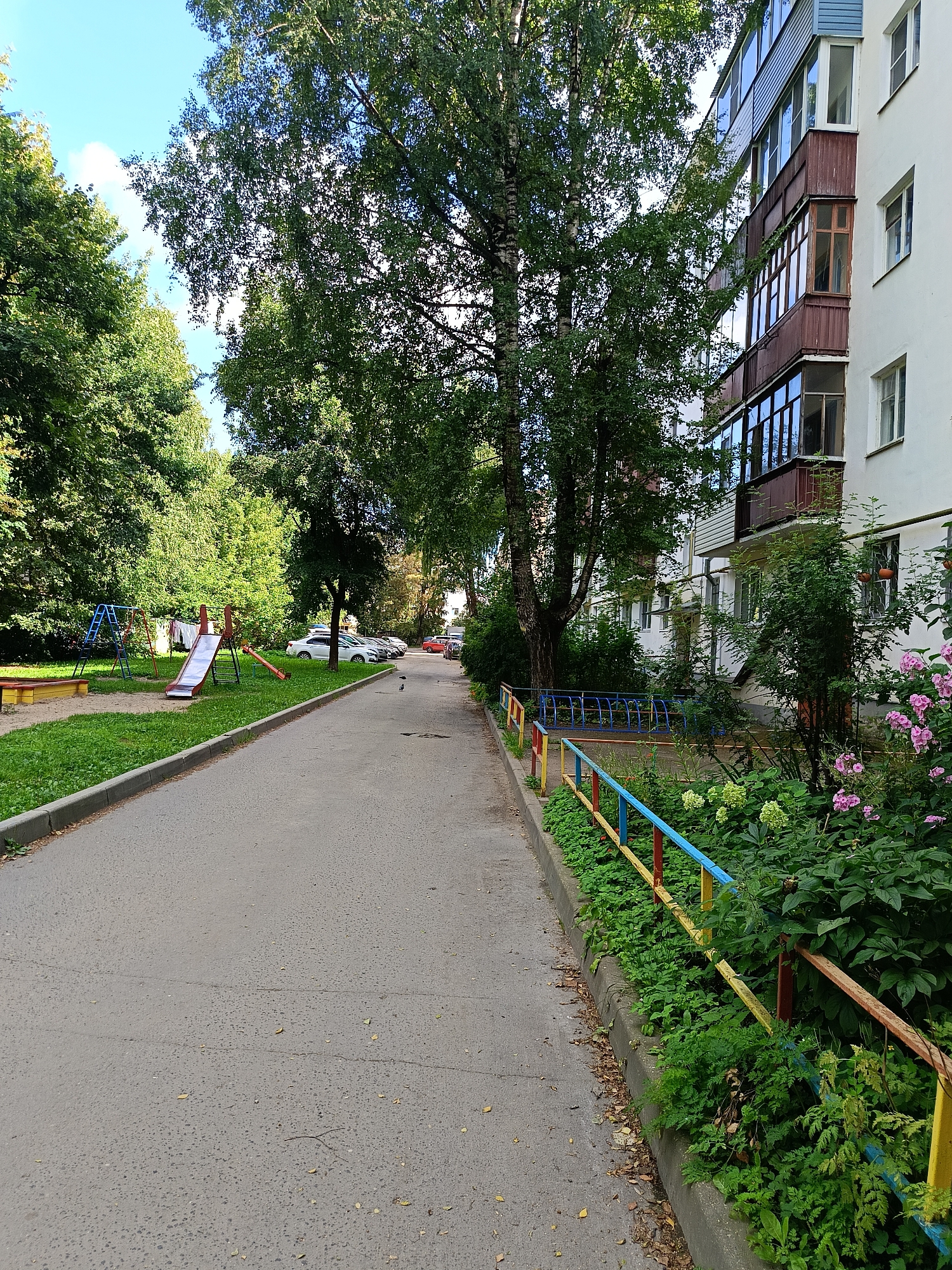 The image shows a view down a paved pathway in a residential area. On the left side of the pathway, there is a playground with a sandbox, swings, a slide, and a seesaw. Beyond the playground, there are several trees and a white car parked. On the right side of the pathway, there is a multi-story building with balconies. The building is white with brown and blue accents. A colorful metal fence, painted yellow, blue, and red, runs along the edge of the pathway and separates it from a green, overgrown area with pink flowers. The sky is blue with some white clouds. A few birds are visible on the pathway in the middle distance.