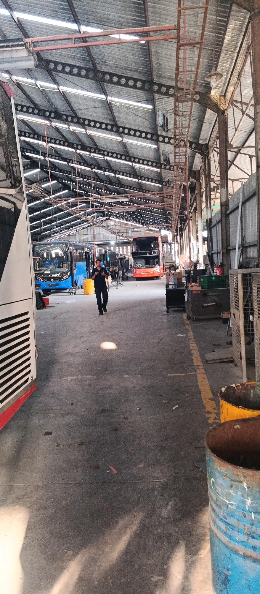 The image depicts the interior of a bus maintenance workshop in Banjararum, Indonesia. A worker in dark clothing walks away from the camera down a concrete aisle. The aisle is flanked by buses, with a white bus on the left and an orange double-decker bus in the background. Various maintenance equipment, tools, and storage containers are visible throughout the workshop. The roof of the workshop is constructed of corrugated metal and exposed steel beams, with natural light entering through gaps and possibly skylights. The overall environment appears industrial and functional, with a focus on vehicle repair and maintenance.
