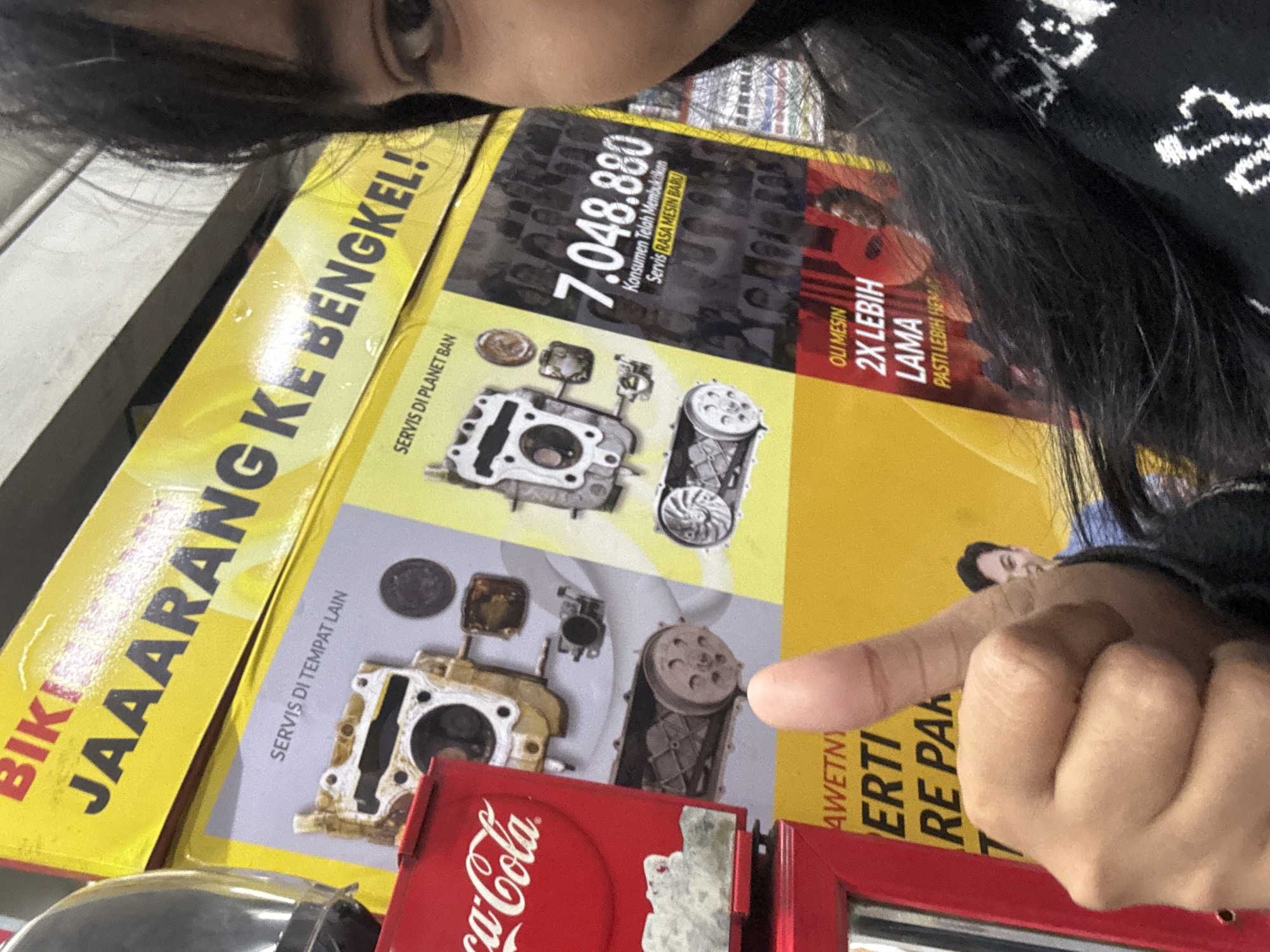 A young woman with dark hair is partially visible in the right side of the frame, looking towards the camera. She is wearing a black sweater with white patterned detailing. Her right hand, with thumb extended upwards in a "thumbs up" gesture, is prominently in the lower left portion of the image. Behind her is a yellow advertisement with bold black text. The text reads "BIKI JAAARANG KE BENGKEL!" and below it, smaller text says "SERVIS DI TEMPAT LAIN" and "SERVIS DI PLANET BAN". To the right of these messages is a graphic displaying the number "7.048.880" followed by text that appears to be in Indonesian. Further down on the yellow advertisement, there is text that reads "OLI MESIN 2X LEBIH LAMA PASTI LEBIH HEMAT". To the left of the woman and the advertisement is a red Coca-Cola branded vending machine. The environment appears to be indoors, possibly a shop or service center. Based on the advertising content related to vehicle servicing and the presence of a vending machine, this scene could be located at a Planet Ban outlet in North Jakarta, Indonesia.