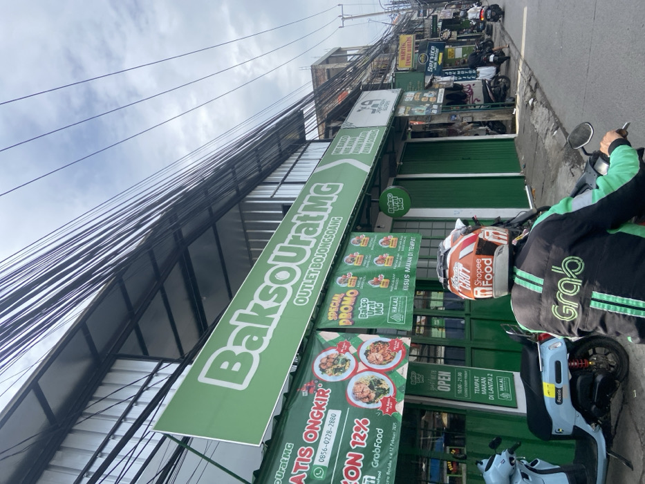The image shows a street scene in Bojongsoang, Indonesia. A motorcyclist wearing a green helmet and black jacket with "Shopee Food" and "Grab" logos stands in front of a storefront with a large green sign that reads "Bakso Urat MG Outlet Bojongsoang" in white letters. There is another sign below that says "Special Promo Bakso Urat MG" in a green box. There are various other signs and objects on the storefront, including "Open 10:00-21:00" and "Tempat Makan Di Lantai 2 Halal." There are two other motorcycles parked in the street. The street has an asphalt surface, and there are many electrical wires overhead. The sky is partly cloudy. The location is a small town in the Bandung Regency, West Java, Indonesia.