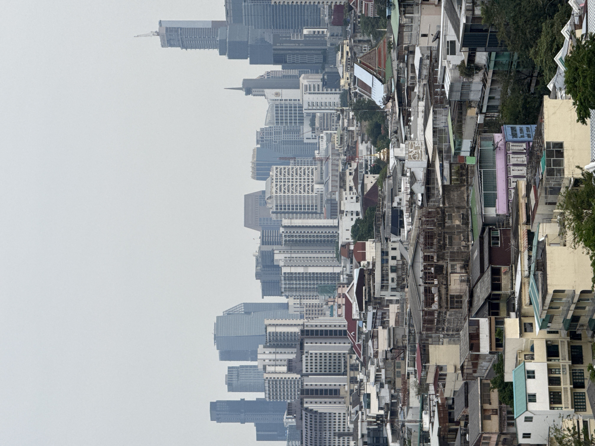 The image depicts a view of Bangkok, Thailand, composed of a mixture of high-rise buildings and residential dwellings in overcast weather. The left side of the image shows a skyline of modern high-rise buildings shrouded in a hazy atmosphere. The right side of the image presents a dense network of lower-rise residential buildings with varied architectural styles and colors. There are no people visible. The environment consists of urban architecture and some trees interspersed within the residential areas.