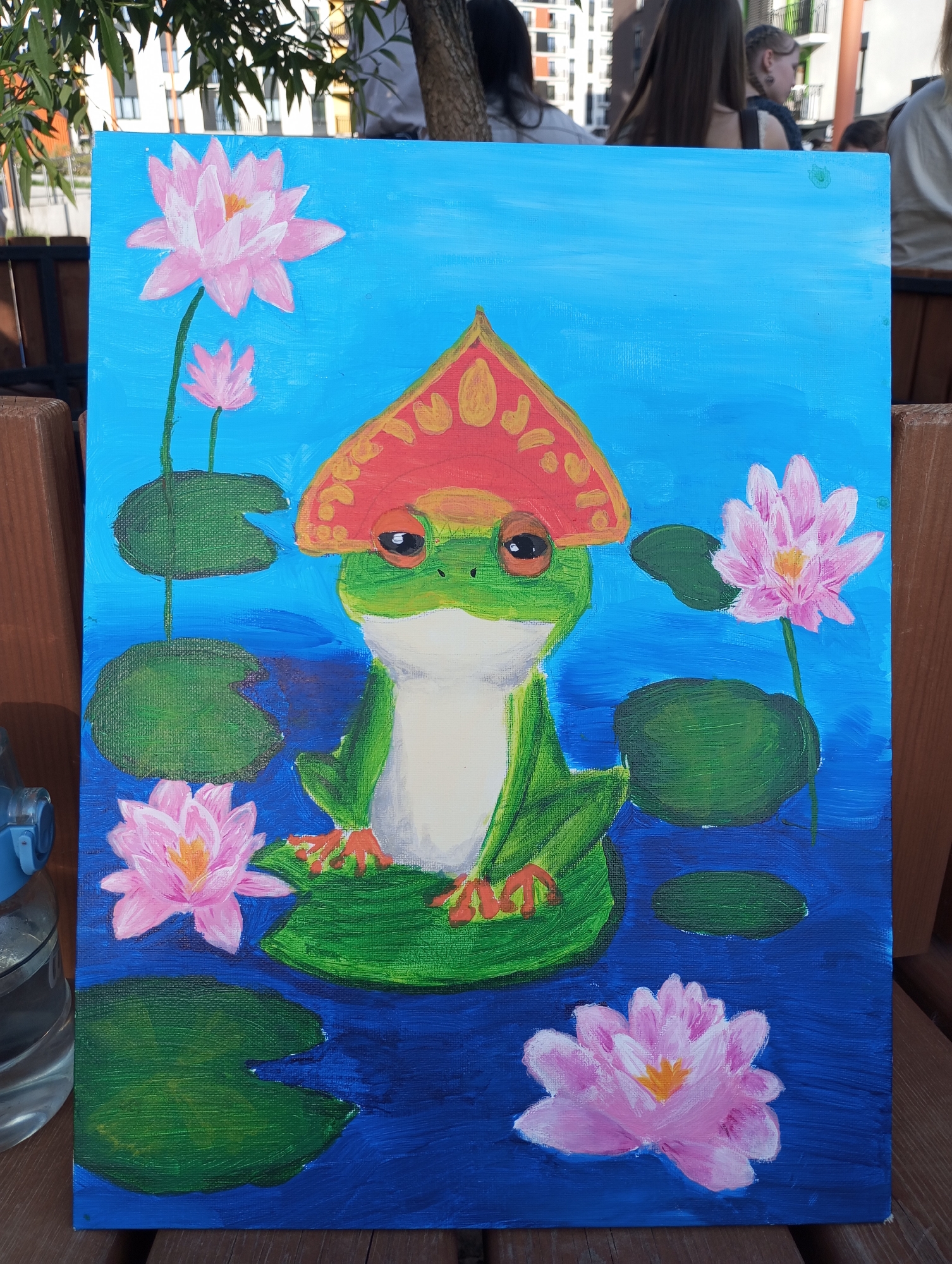 A painting is displayed outdoors, resting on a wooden surface. The painting features a green frog wearing a red hat with yellow details. The frog sits on a lily pad, surrounded by pink water lilies and additional lily pads on a blue background. Other people are partially visible in the background, along with parts of buildings and a tree. The location is possibly the city of Pervomaysky, Russia.