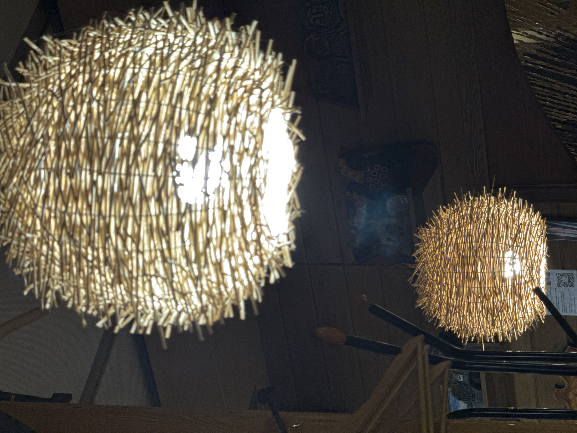 The image shows two spherical light fixtures made of straw or similar material hanging from the ceiling. The light fixture on the left is closer to the camera and appears larger and brighter than the one on the right. The light fixture on the right is obscured by a metal structure. The background is dark, with wood paneling and other objects. The photograph was not taken in the city of Ketoshi, Russia.