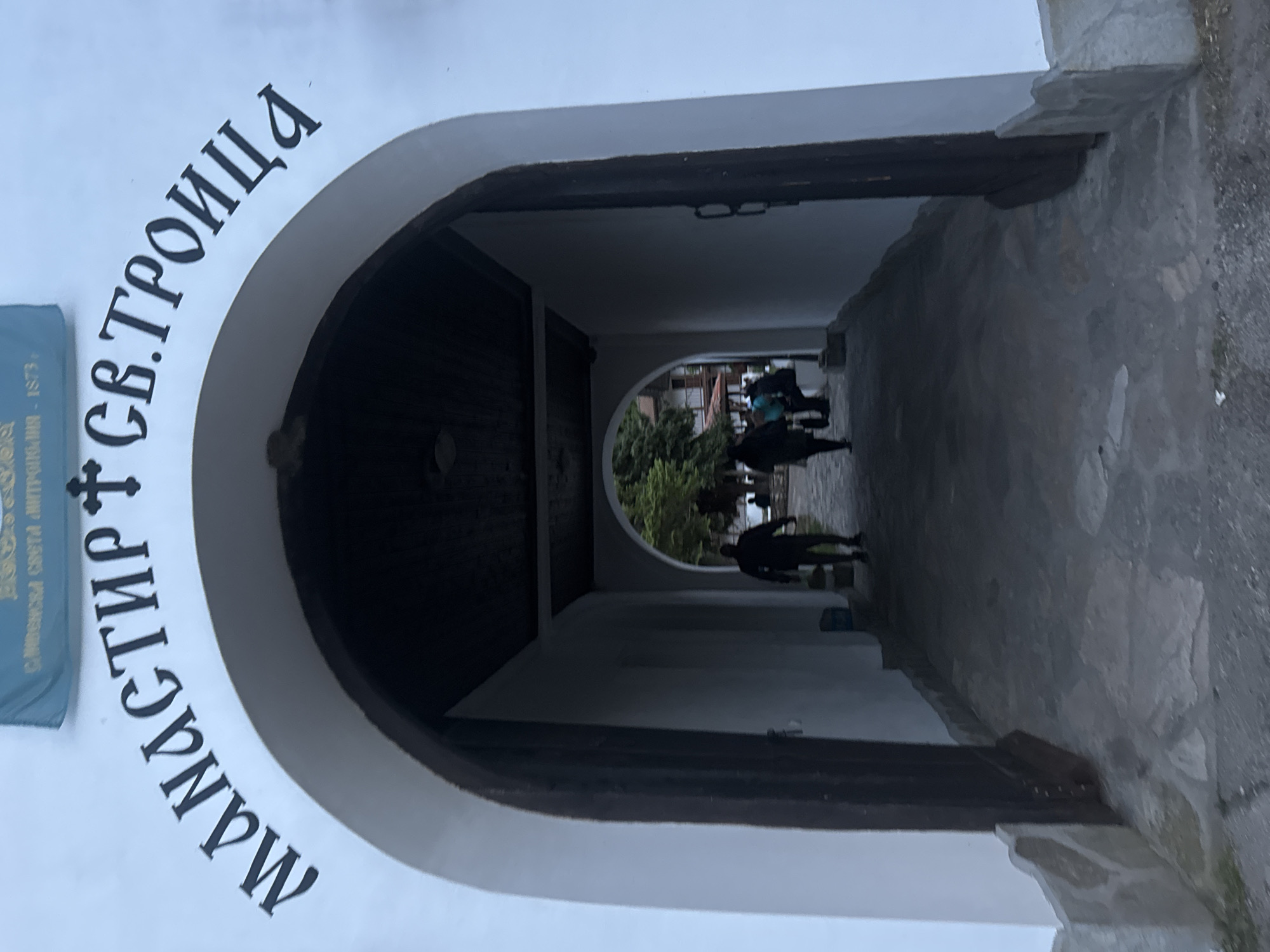 The image depicts the entrance to the Holy Trinity Monastery in the village of Mramor, Pernik Province, Bulgaria. The entrance is an arched opening in a white wall with the monastery's name written in Cyrillic lettering above the arch: "Mанастир Св. Троица" ("Monastery of the Holy Trinity"). The archway leads to a dimly lit corridor with another archway at the far end. People are visible walking through the corridor.