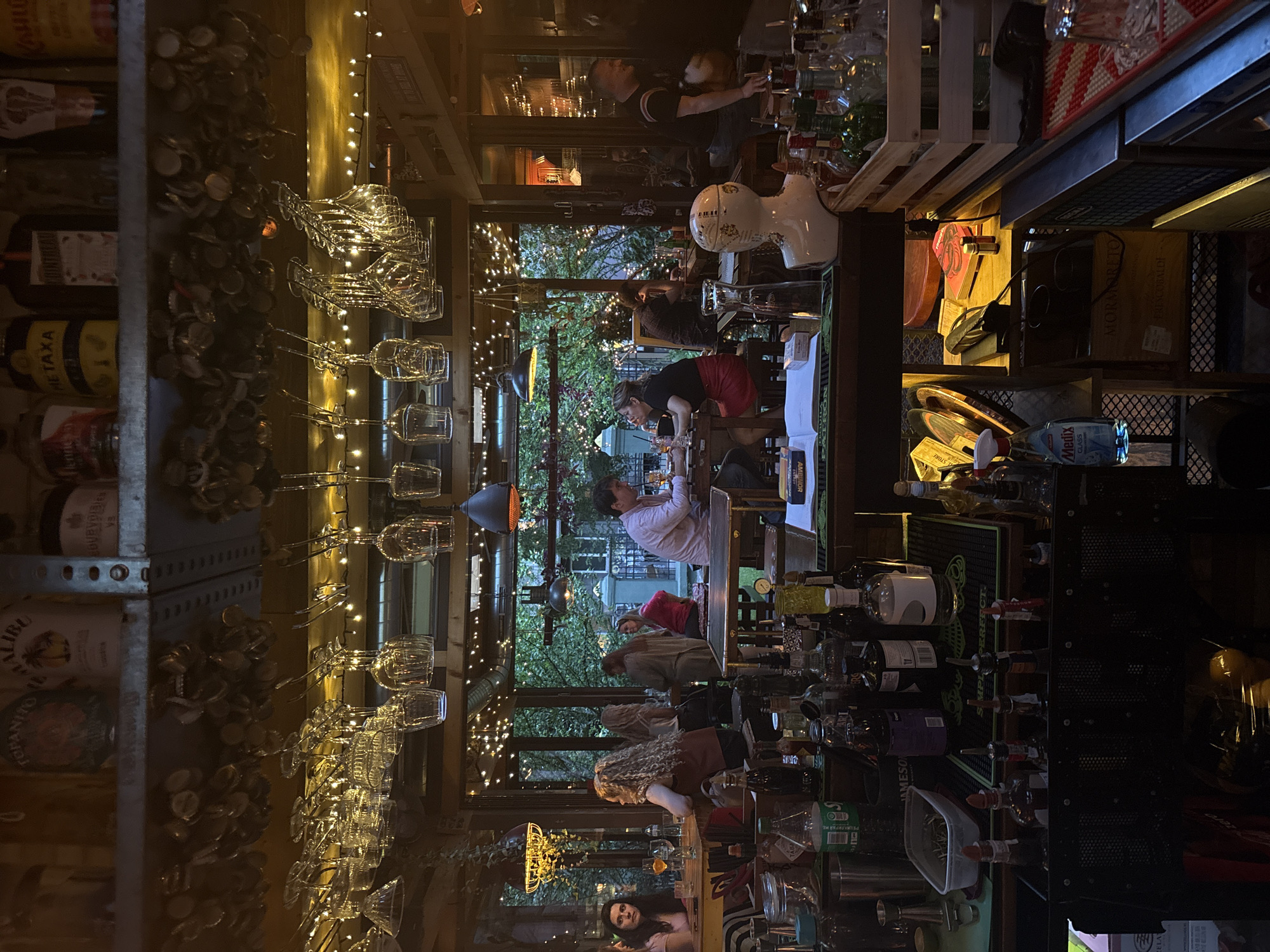 This media file captures a vibrant evening scene inside a bar or restaurant in Sofia, Bulgaria, looking out onto an illuminated outdoor patio area. The setting is warm and inviting, with numerous string lights adding to the ambiance both indoors and through the windows, suggesting it is nighttime.
Several people are present, engaging in various activities. Indoors, a bartender is visible behind the counter, preparing drinks. Other patrons are seated at tables or along the bar, socializing and looking at menus or phones. Outdoors, more individuals are seated at tables in a garden or patio space, also appearing to be conversing and enjoying the evening.
Notable objects within the bar include shelves heavily decorated with a collection of bottle caps, and rows of clear wine glasses hanging upside down. Various bottles of spirits and wines are displayed, some identifiable by their labels: "MALIBU," "METAXA," "JAMESON," "TOPANITO D," "BOURVOLEE Vs," "AMBINOR," and "THOLST." On the bar counter, a mat with "PERFECT." is visible, alongside other bottles and bar equipment, including a distinctive white decorative head. A "Medix GLASS" bottle and a box labeled "MORMORETO FRESCOBALDI" with "KAHLE" are also discernible. The overall atmosphere conveys a lively and relaxed social gathering.
