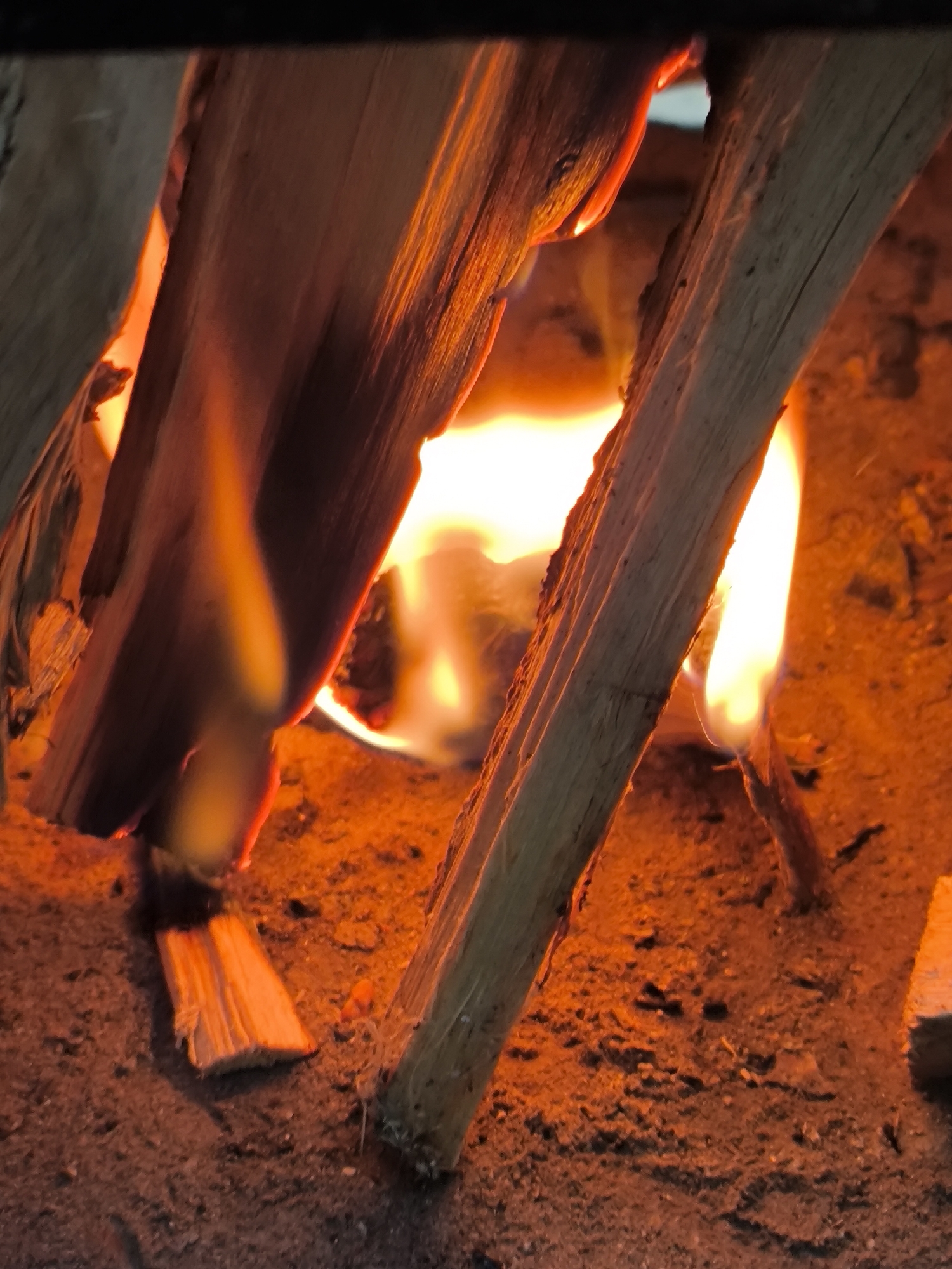 The image is a close-up shot of burning logs in a fire pit. The logs are made of light-colored wood and are arranged in a triangular formation, with flames visible between them. The flames are bright orange and yellow, casting a warm glow on the surrounding ash and embers. The background is dark and out of focus, suggesting the photograph was taken in low light conditions. There are no people or other discernible objects in the frame. The scene appears to be outdoors, given the presence of ash and dirt. There is no information in the image to indicate it is located in Новоникольское, Russia.