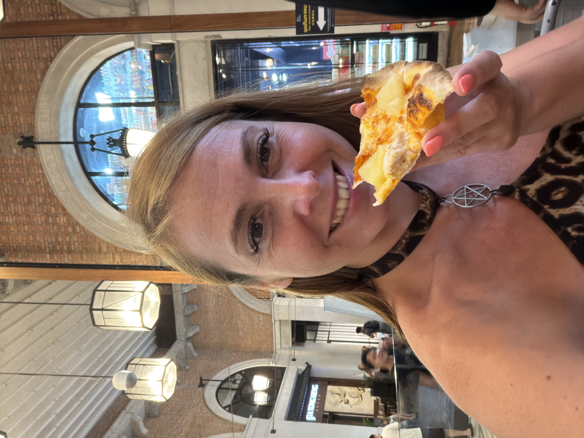 A woman with blonde hair and light skin is taking a selfie while holding a slice of pizza. She is smiling broadly and has makeup on, including eyeliner and mascara. She is wearing a leopard-print top and a black choker with a silver pentagram pendant. The background shows an indoor environment with brick walls, arched windows, and hanging lanterns. In the distance, there are other people and storefronts, suggesting a commercial area or marketplace. One storefront sign reads "JIATONGHENG" in English. There is also Thai text visible on a sign to the right, indicating the location is likely in Thailand. The overall impression is a casual, personal moment captured in a public space.