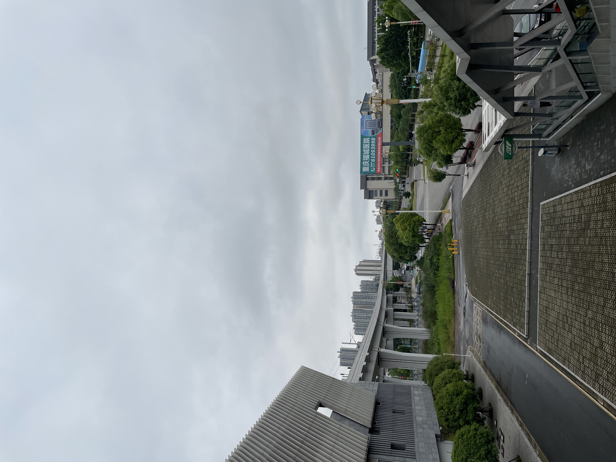 The view is from an elevated perspective, looking down a street in the Jiangjin district of Chongqing, China. The sky is overcast. A building with a corrugated roof is in the foreground. A raised highway or elevated train line runs along the right side of the street, with concrete supports and a road passing underneath. Various buildings of varying heights and designs, mostly residential, are visible in the distance. A billboard advertising Chongqing Fucheng Hospital is visible. The street is paved with permeable pavers, and there is a sidewalk bordered by a line of trees. A modern building structure that appears to be an entryway to a building or station is on the right.