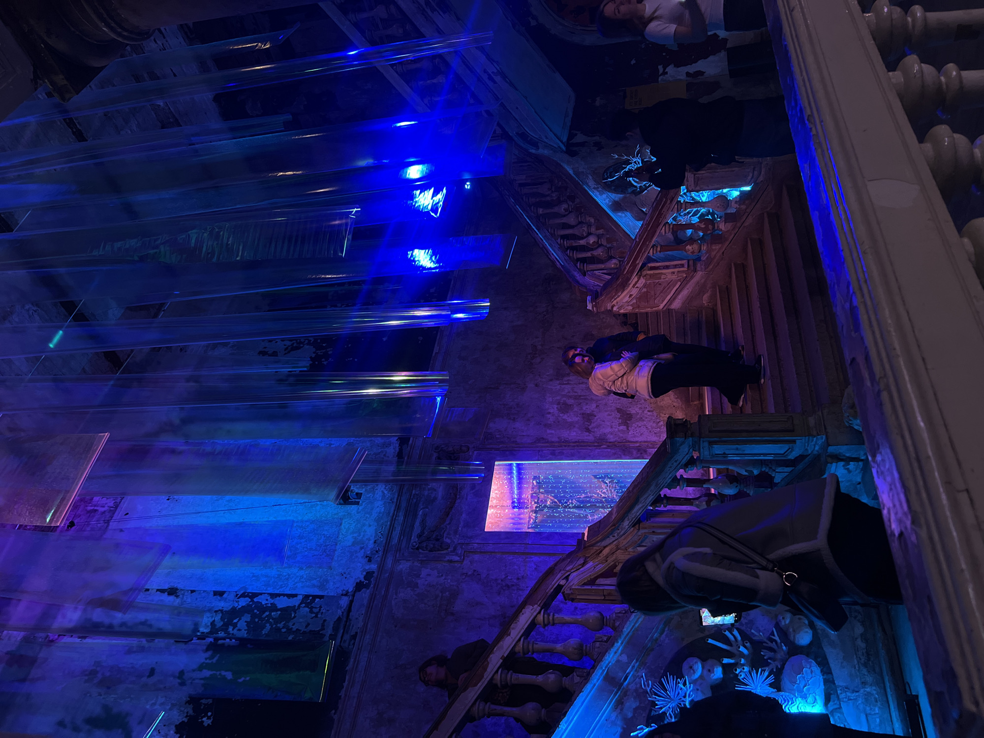 A tilted, low-angle shot captures the interior of a building, possibly located in Saint Petersburg, Russia, due to the architectural style and historical appearance of the walls. The walls are aged, showcasing a distressed texture with areas of peeling paint and visible wear. The scene is bathed in artificial lighting, predominantly in shades of blue, purple, and pink, creating an otherworldly atmosphere. The primary focus is on a staircase, which appears to be of classic design with detailed balusters. People are situated on different levels of the staircase. Hanging from the ceiling are large, transparent panels or tubes, which reflect and refract the artificial lighting, adding a modern artistic element to the historic setting. These panels add depth and complexity to the scene, juxtaposing the old with the new. The image also includes details like fragments of architectural decorations, such as molded details on the walls and around the staircase. The overall impression is a blend of historic architecture and contemporary art installation, creating a unique and visually intriguing scene.