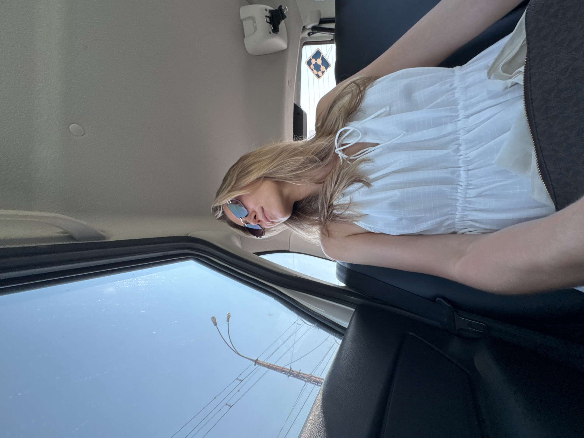 A blonde woman with sunglasses sits in the back of a car, looking out the window. She wears a white sundress and has a dark handbag on her lap. The car's interior is visible, with black seats and the roof lining above. Outside the window, a light pole with wires and a blue sky are visible. A blue and white diamond-shaped sticker is affixed to the rear window.
