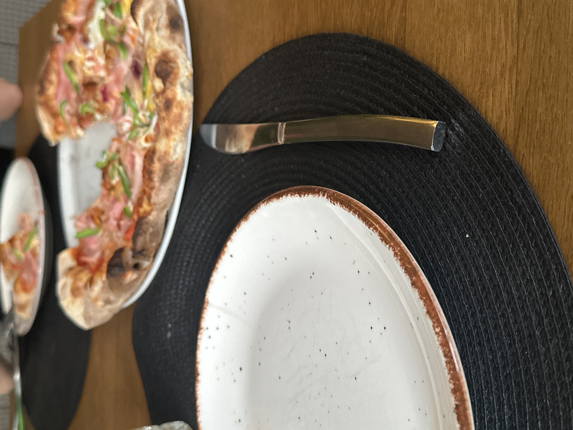 The image shows a table setting with a partially eaten pizza on a white plate to the upper left. Another, smaller white plate with pizza is visible to the left of the main pizza plate. A butter knife with a gold-colored handle is positioned horizontally between the pizza plate and an empty white plate with a speckled pattern and a brown rim, placed on a black woven placemat. The table surface appears to be wooden. The location is not discernable from the image, and therefore it's impossible to determine if it is in Sofia, Bulgaria.